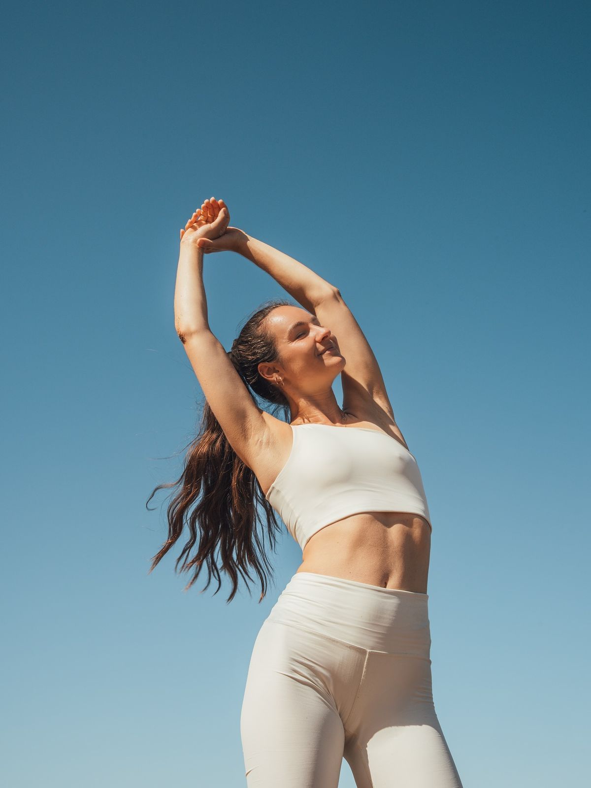 Woman stretching outdoors against a clear blue sky, wearing a white sports bra and leggings.