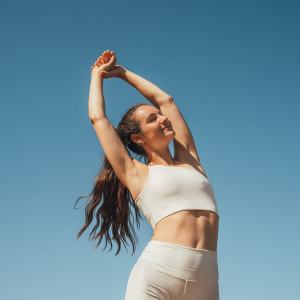 Woman in white activewear stretching with arms overhead against a clear blue sky