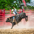 Bull rider on a bucking black bull in a dusty rodeo arena, red fencing and an American flag behind.
