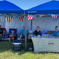 Blue tents at an outdoor event with people seated behind a registration table and patriotic bunting.