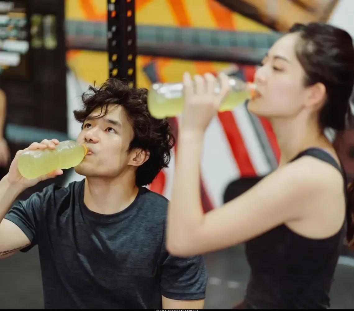 Two people resting in a gym, one drinking from a yellow bottle and the other holding a water bottle