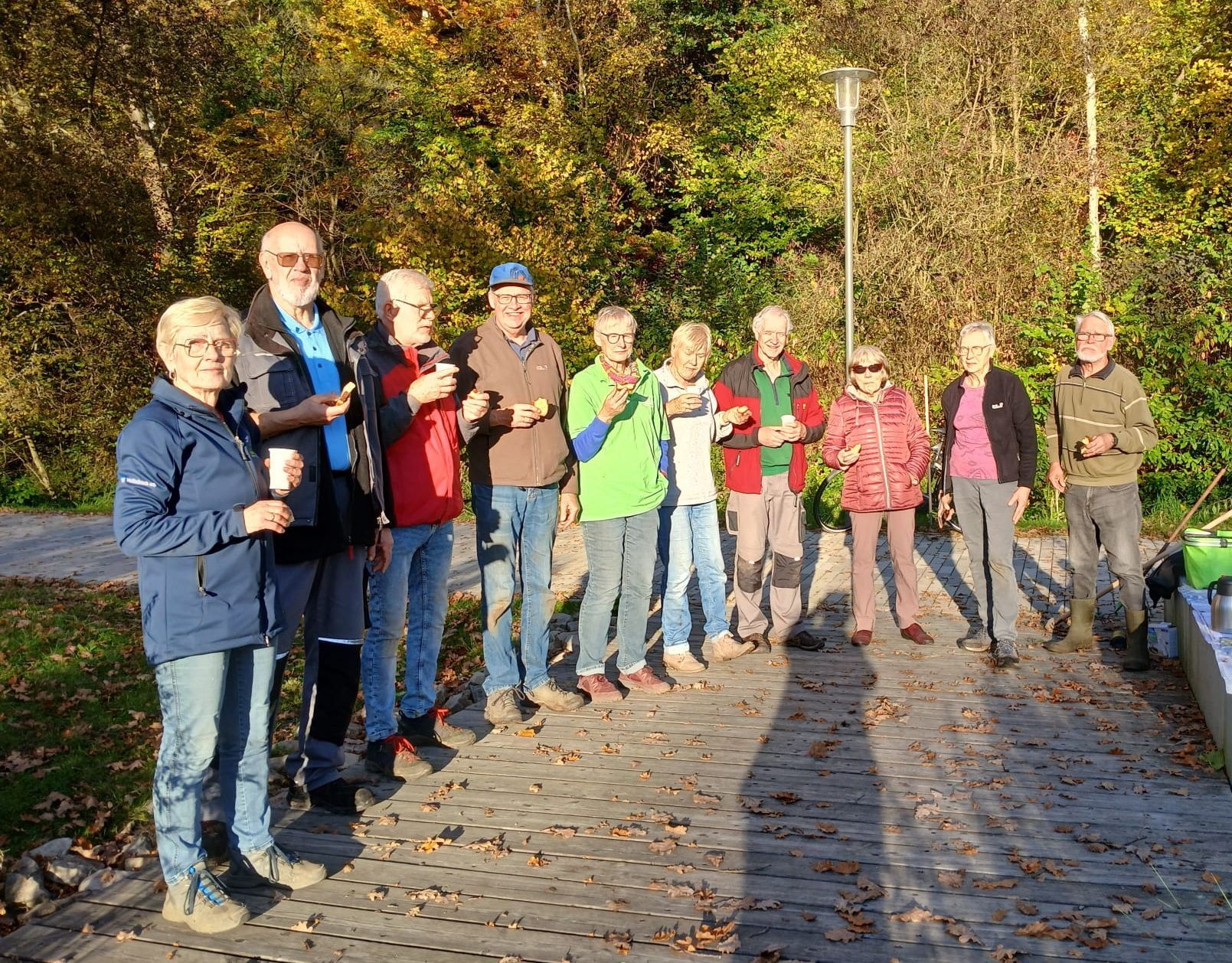 Eine Gruppe von Menschen mit kleinen Bechern steht im Freien auf einem gepflasterten Weg, umgeben von Bäumen.