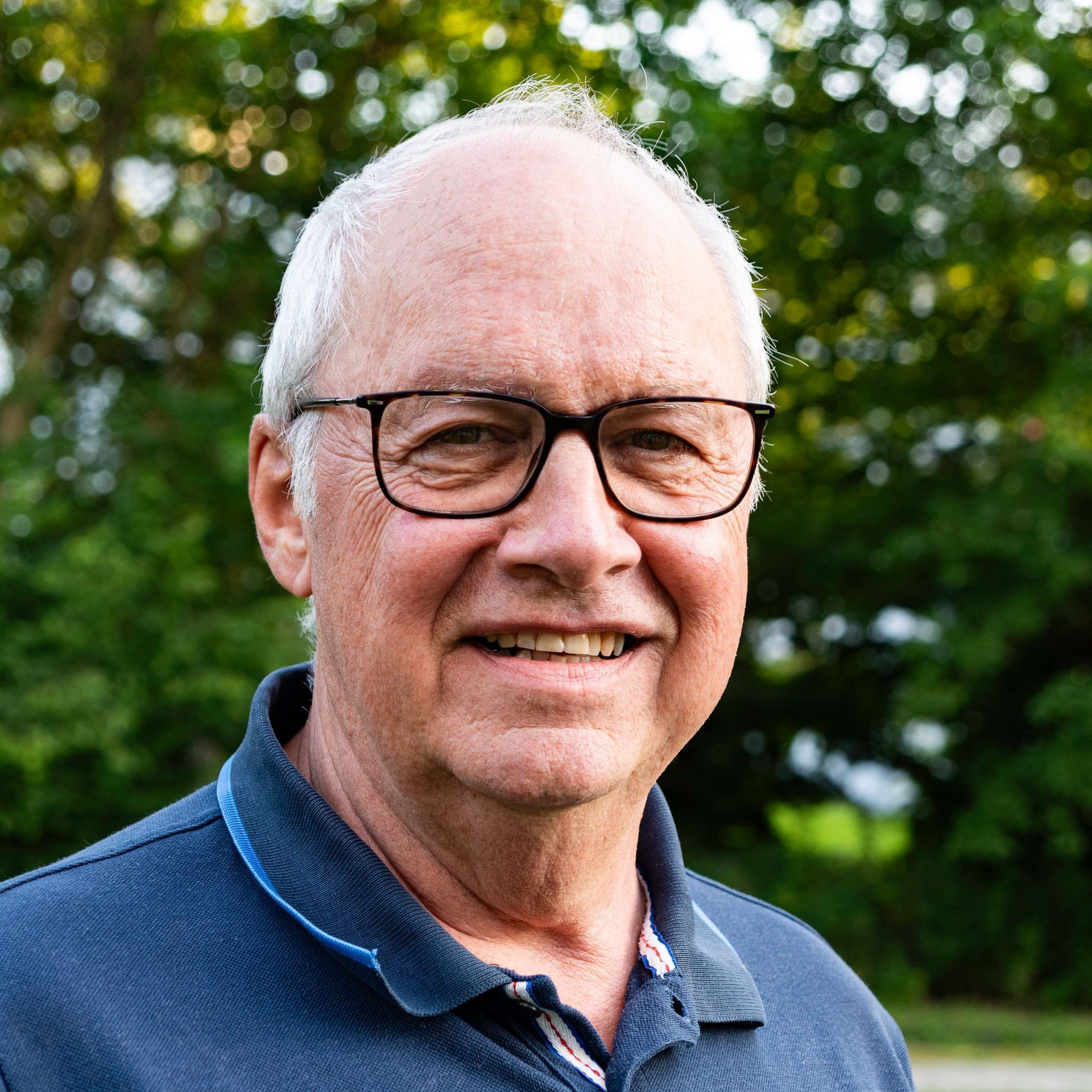 Mann mit Brille, lächelnd, trägt ein blaues Poloshirt im Freien vor verschwommenem grünem Hintergrund.