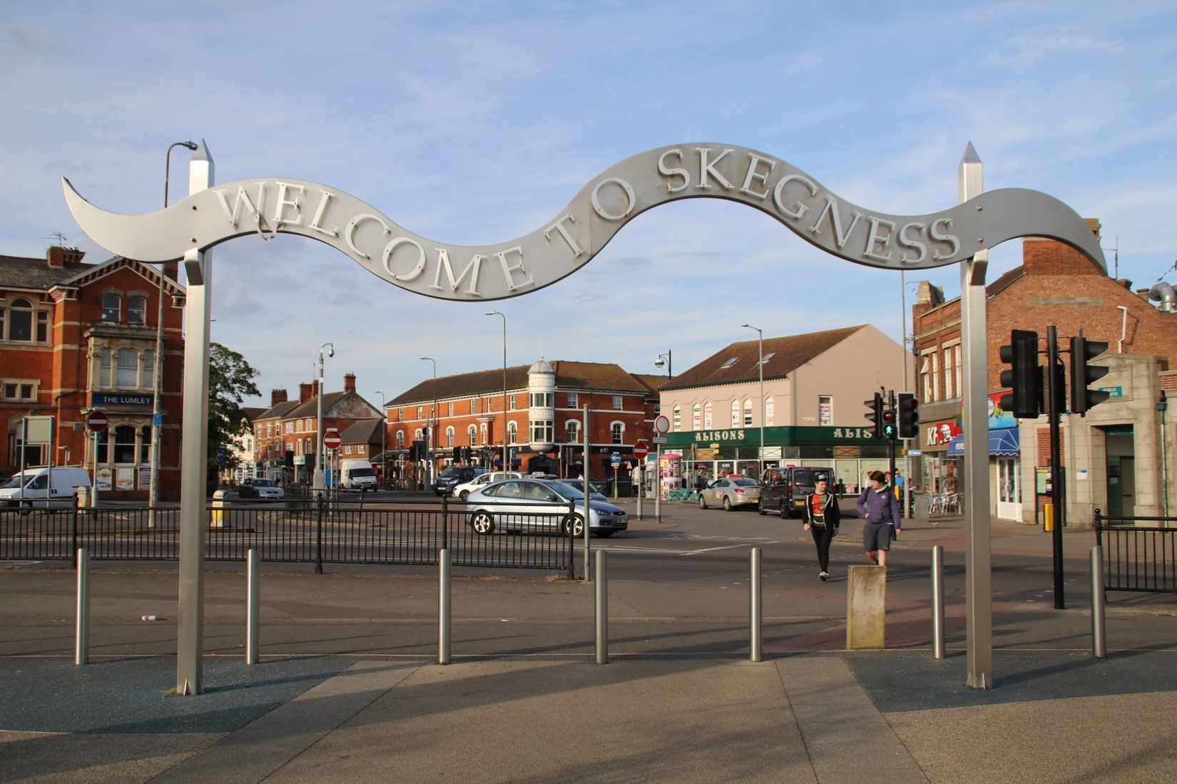 Ein Schild mit der Aufschrift „Willkommen in Skegness“ wölbt sich über einen Stadtplatz mit Gebäuden, Autos und Menschen.
