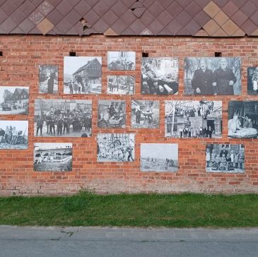 Schwarz-Weiß-Fotos sind an einer Backsteinmauer angebracht. Die Fotos zeigen Gruppen von Menschen und Szenen aus einem Dorf.