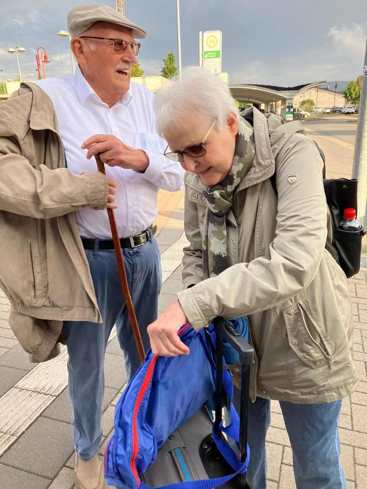 Ein älteres Ehepaar an einer Bushaltestelle. Einer hält einen Gehstock, der andere schaut in einen blauen Rucksack. Bewölkter Tag.