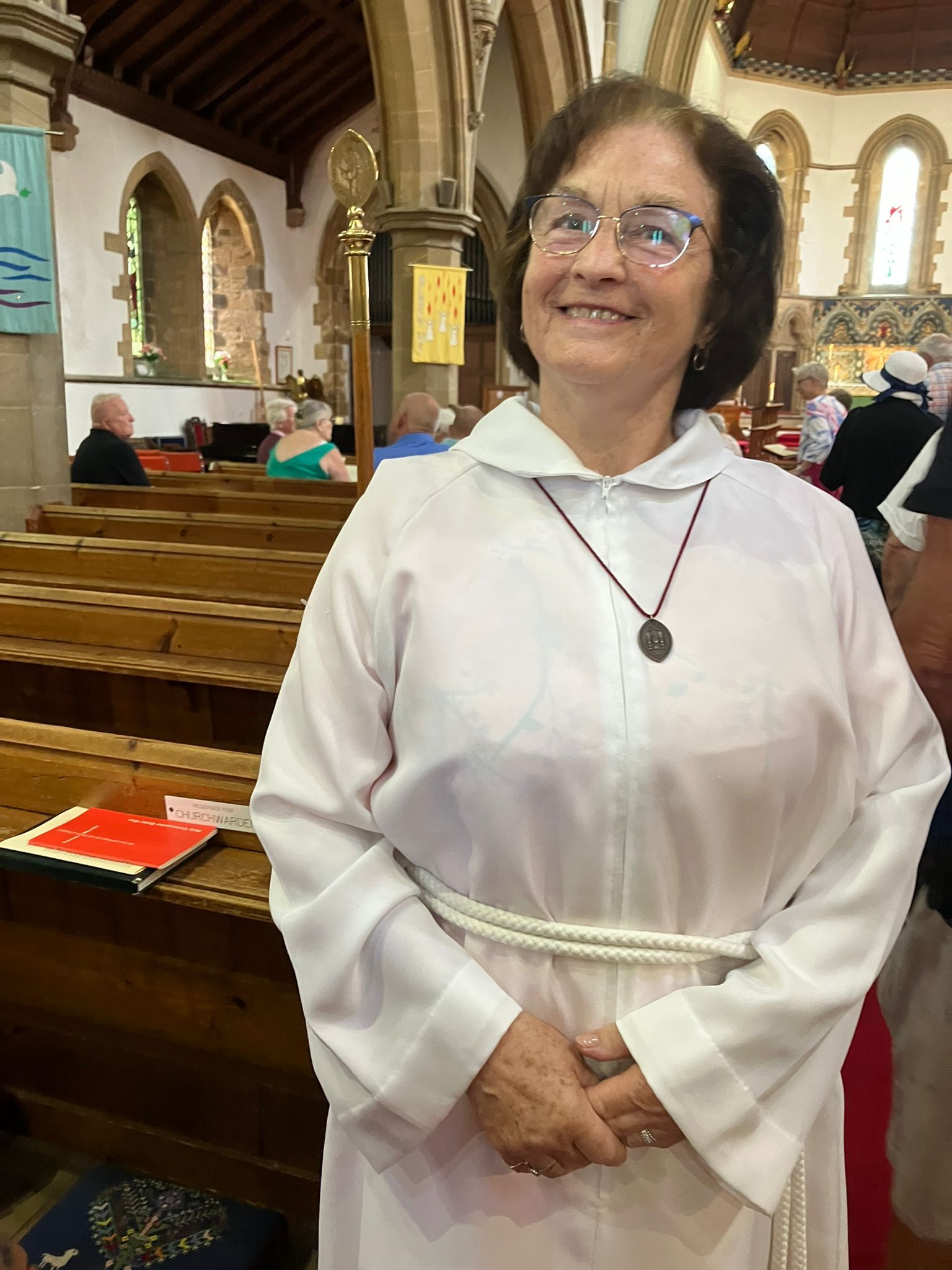 Eine Frau in weißem Gewand und mit Brille lächelt in einer Kirche, in der Nähe der Kirchenbänke.
