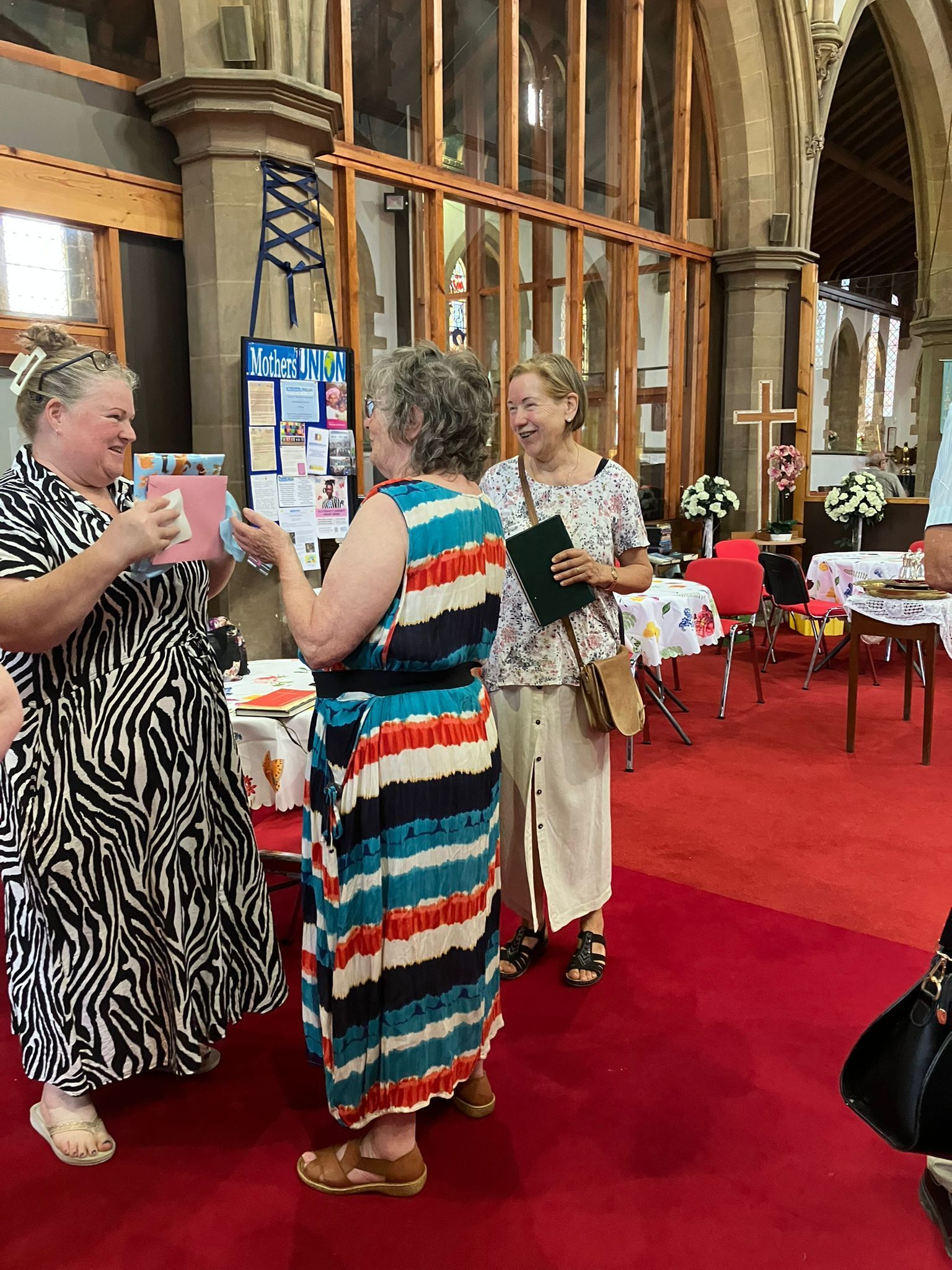 Drei Frauen in einer Kirche, lächelnd und sich unterhaltend. Eine hält ein Geschenk, im Hintergrund ein roter Teppich und Blumen.