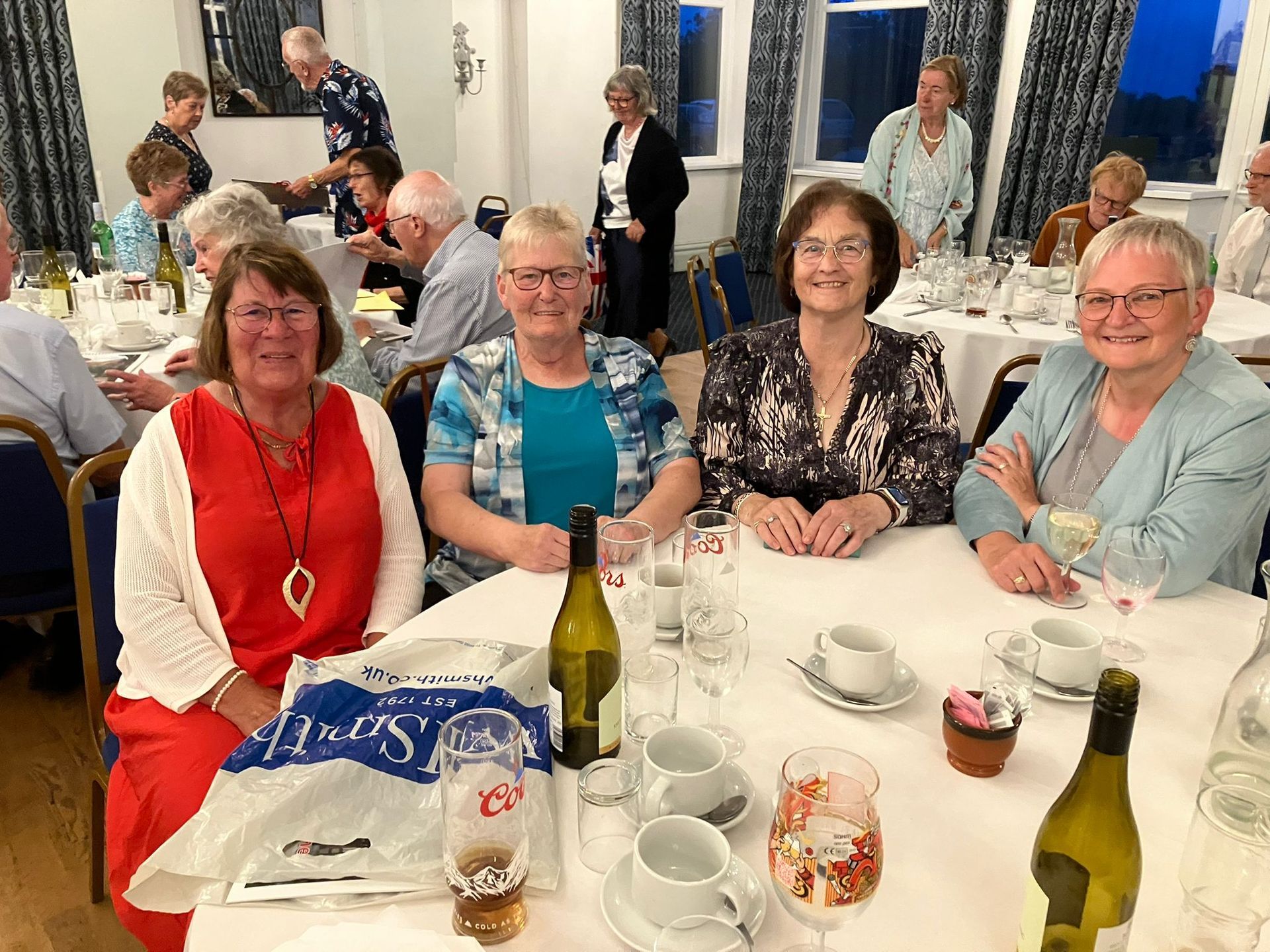 Vier Frauen lächeln an einem zum Abendessen gedeckten Tisch. Weitere Personen und Tische befinden sich in einem hell erleuchteten Raum.