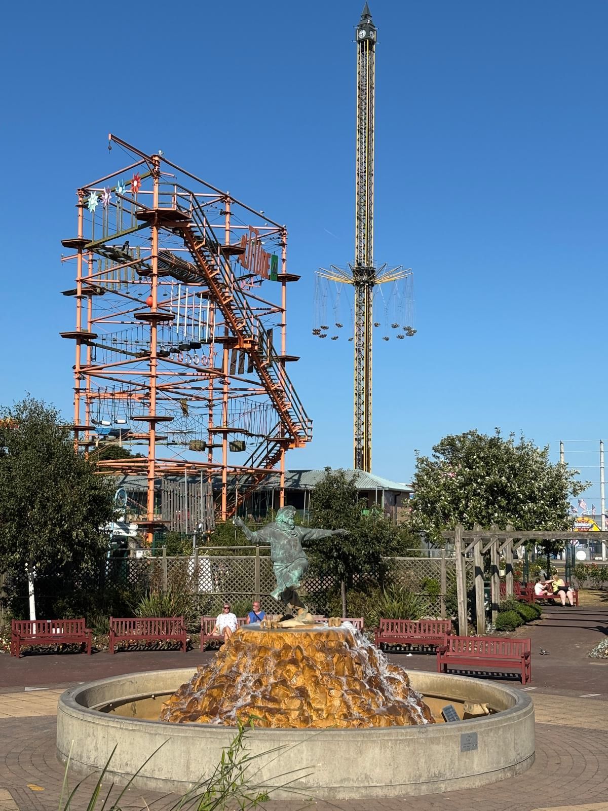 Ein Vergnügungspark mit Hochseilgarten, Freifallturm und Springbrunnen unter blauem Himmel.