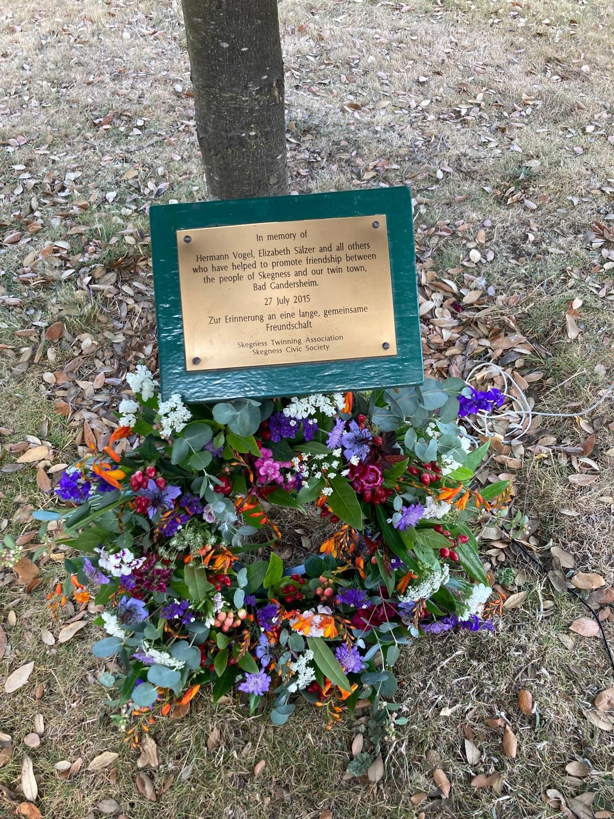 Ein Gedenkkranz mit bunten Blumen liegt unterhalb einer grünen Gedenktafel und einem Baumstamm, der in einem Feld aufgestellt ist.