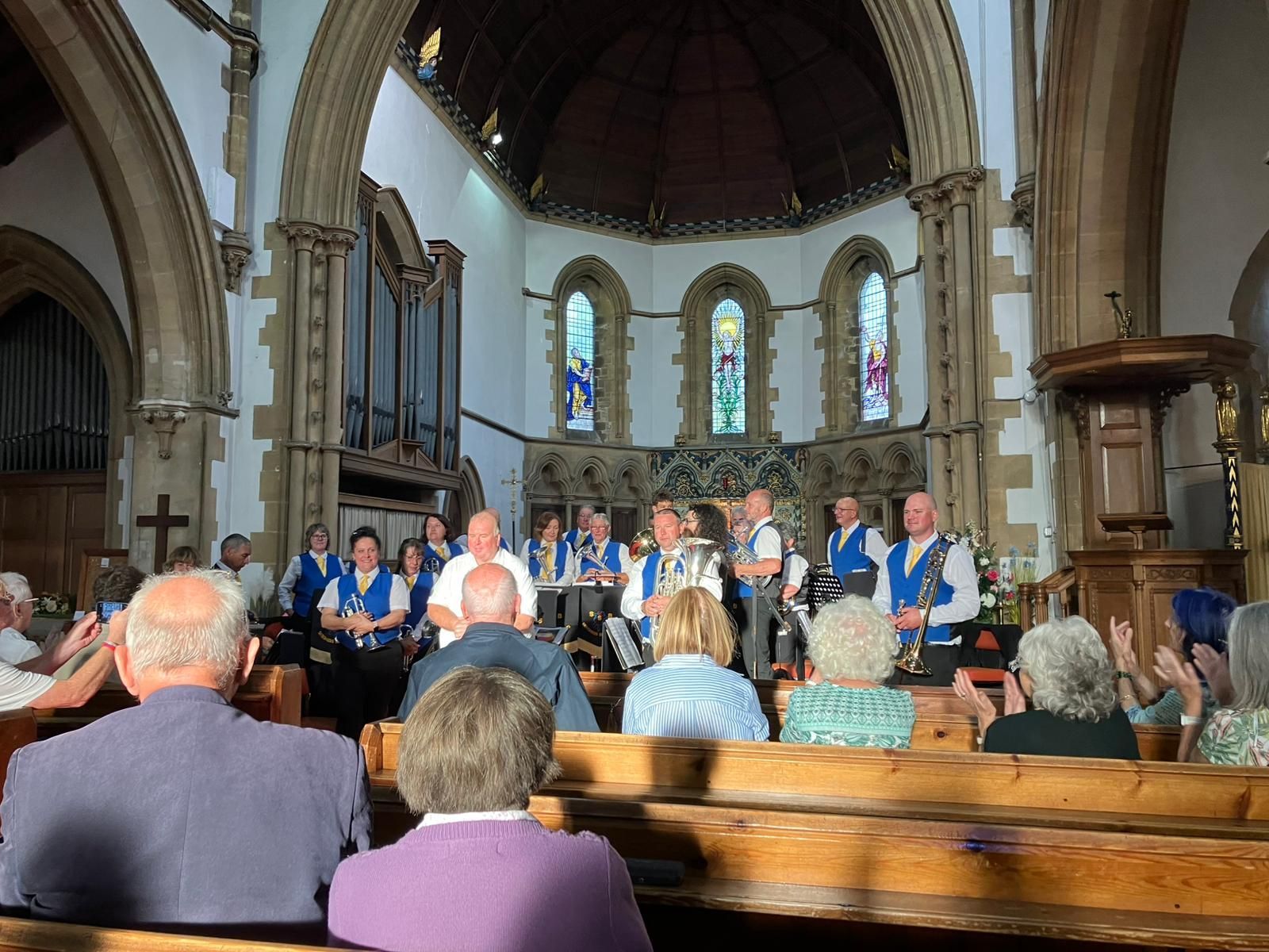 Eine Band spielt in einer Kirche, das Publikum klatscht. Die Musiker tragen Blau. Licht strömt durch die Buntglasfenster.