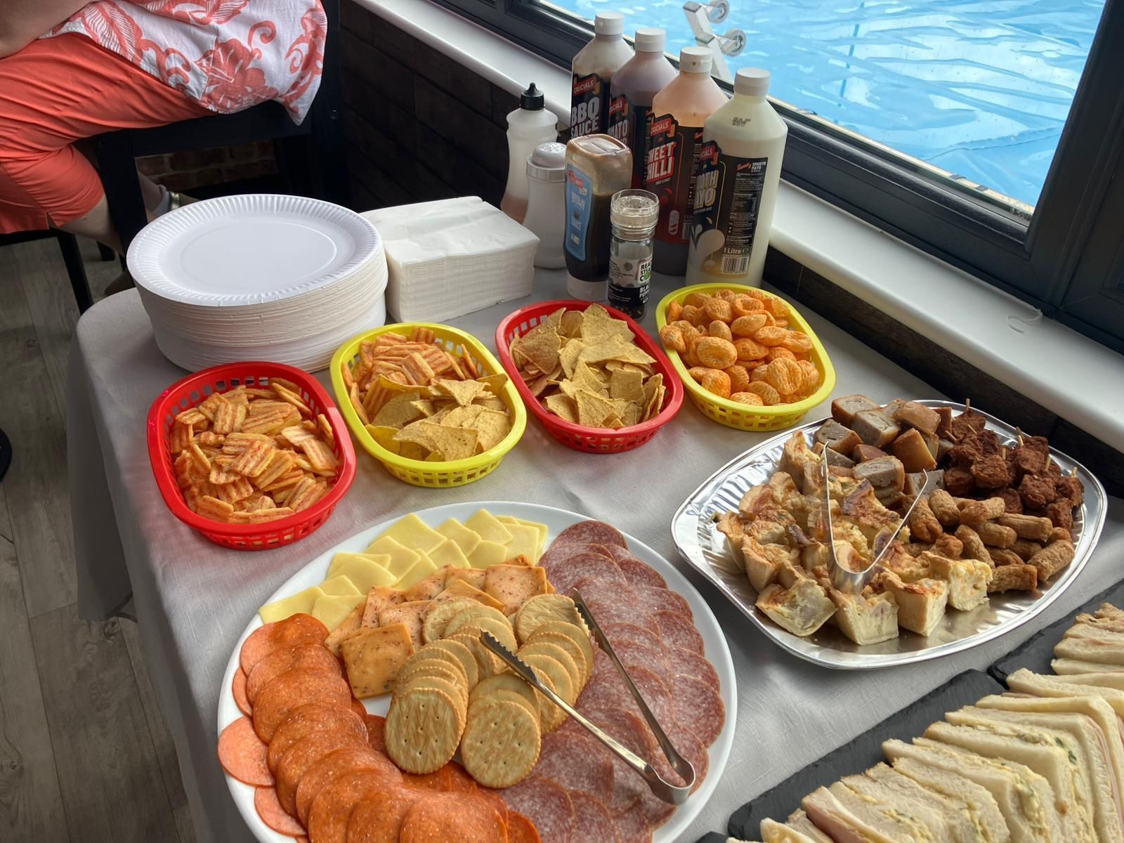 Ein Tisch mit verschiedenen Snacks: Chips, Käse, Wurstwaren und Sandwiches. Getränke und Teller sind ebenfalls vorhanden.
