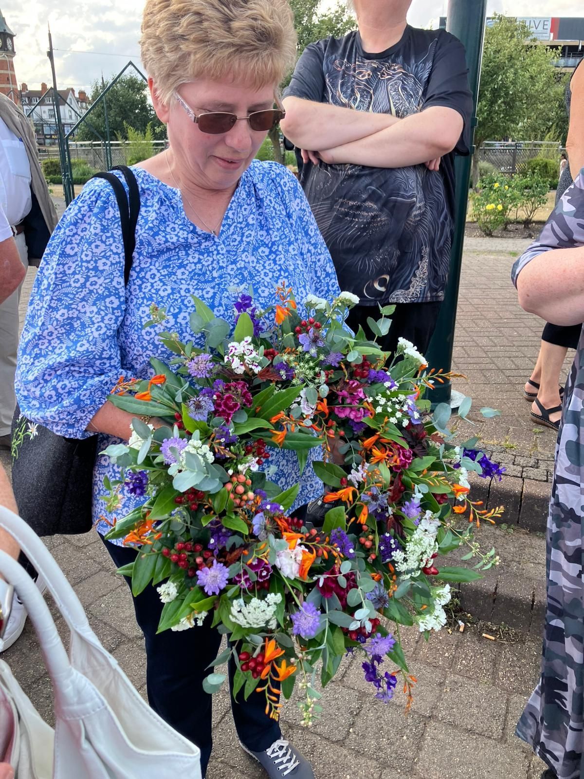 Eine Frau hält im Freien einen großen Kranz aus bunten Blumen. Umstehende beobachten sie.
