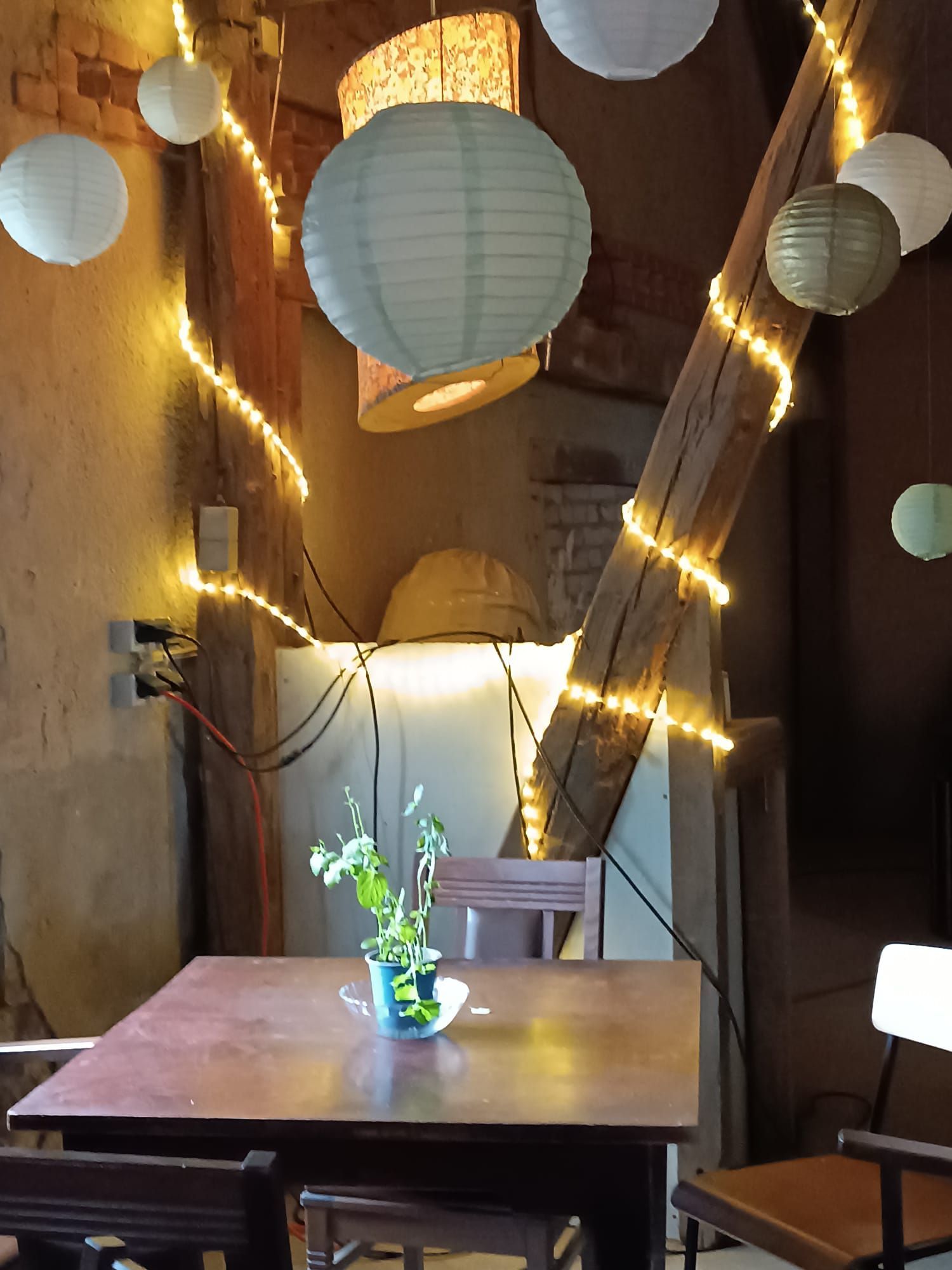 Ein Holztisch und Stühle in einem warm beleuchteten Raum mit Papierlaternen und Lichterketten, ein kleines Blumenarrangement auf dem Tisch.