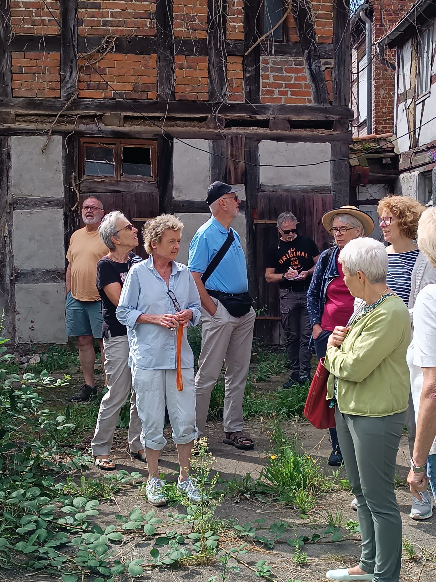 Eine Gruppe von Menschen vor einem Gebäude aus Ziegeln und Holz hört einer Person zu, die einen orangefarbenen Stock hält.
