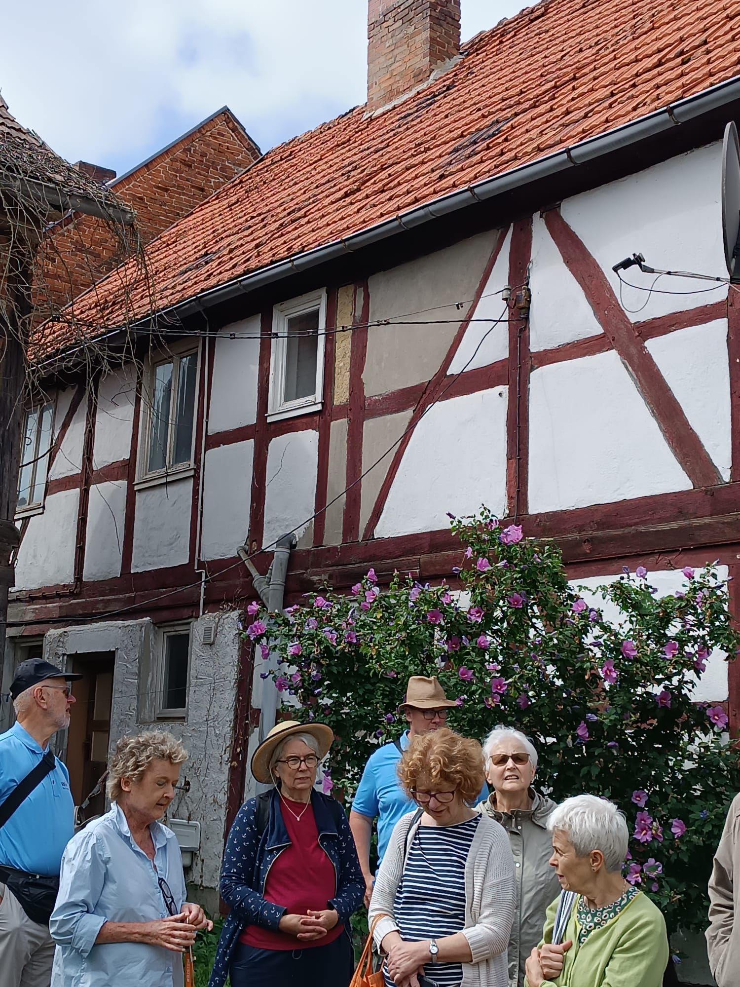 Eine Gruppe von Menschen vor einem Fachwerkhaus mit rotem Ziegeldach und blühenden Büschen.