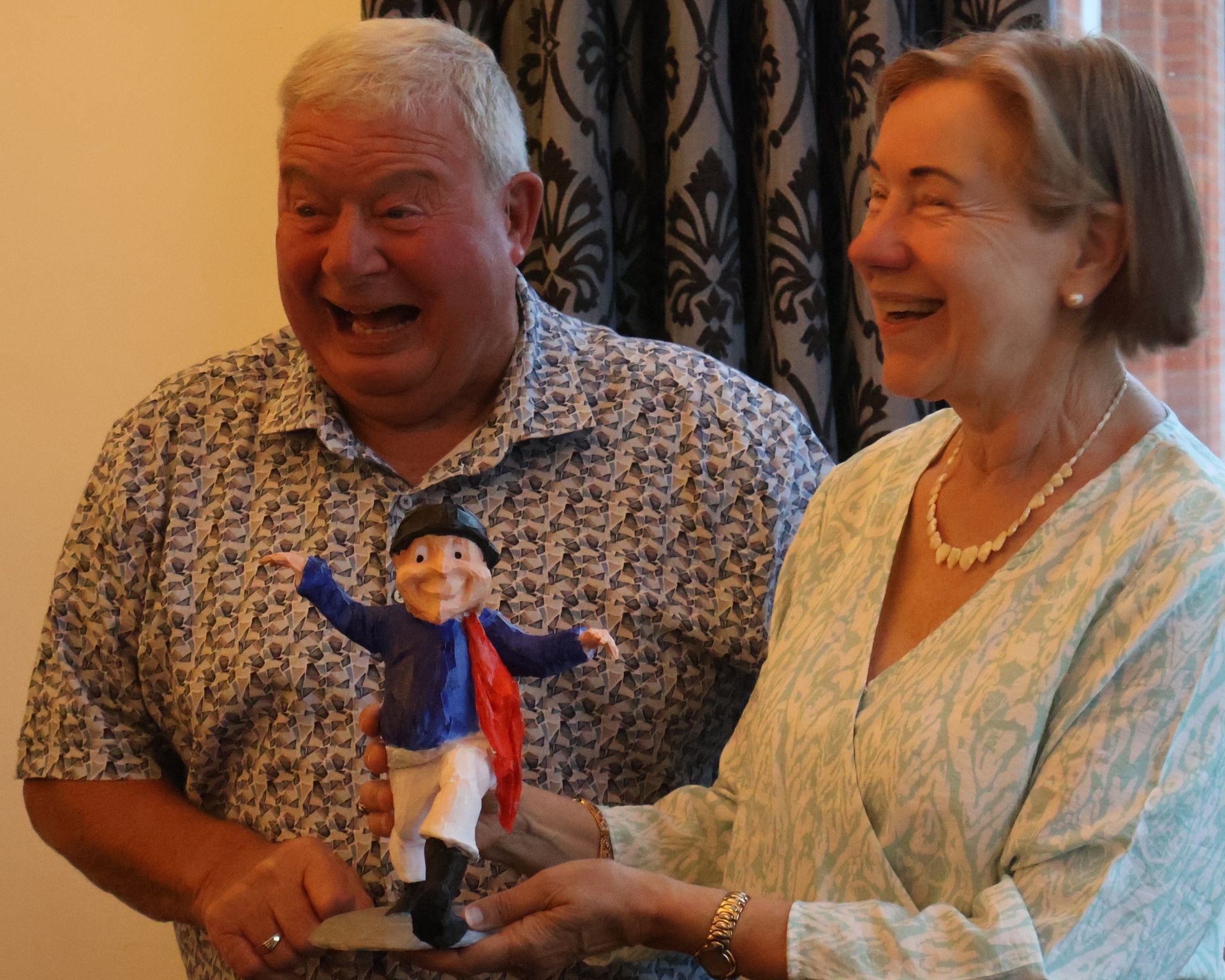 Ein Mann und eine Frau halten aufgeregt eine kleine Figur und lachen beide. Blaue, weiße und rote Kleidung ist zu sehen.