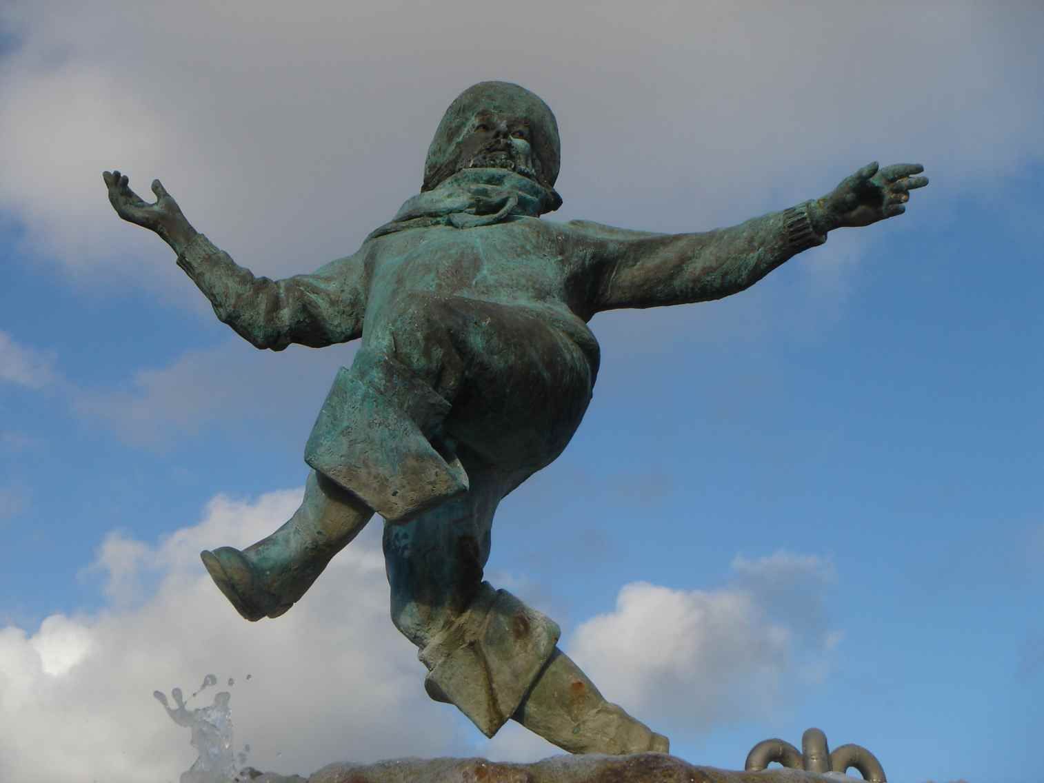 Bronzestatue einer Person in vollem Schritt, die Arme ausgestreckt, vor einem blauen Himmel mit Wolken.