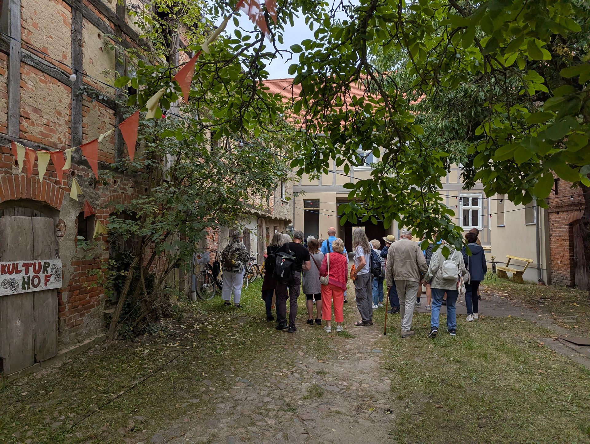 Eine Gruppe von Menschen steht im Freien und betrachtet ein Gebäude. Links ist ein teils aus Holz, teils aus Ziegeln bestehendes Bauwerk zu sehen. Darüber ragen grüne Bäume empor.