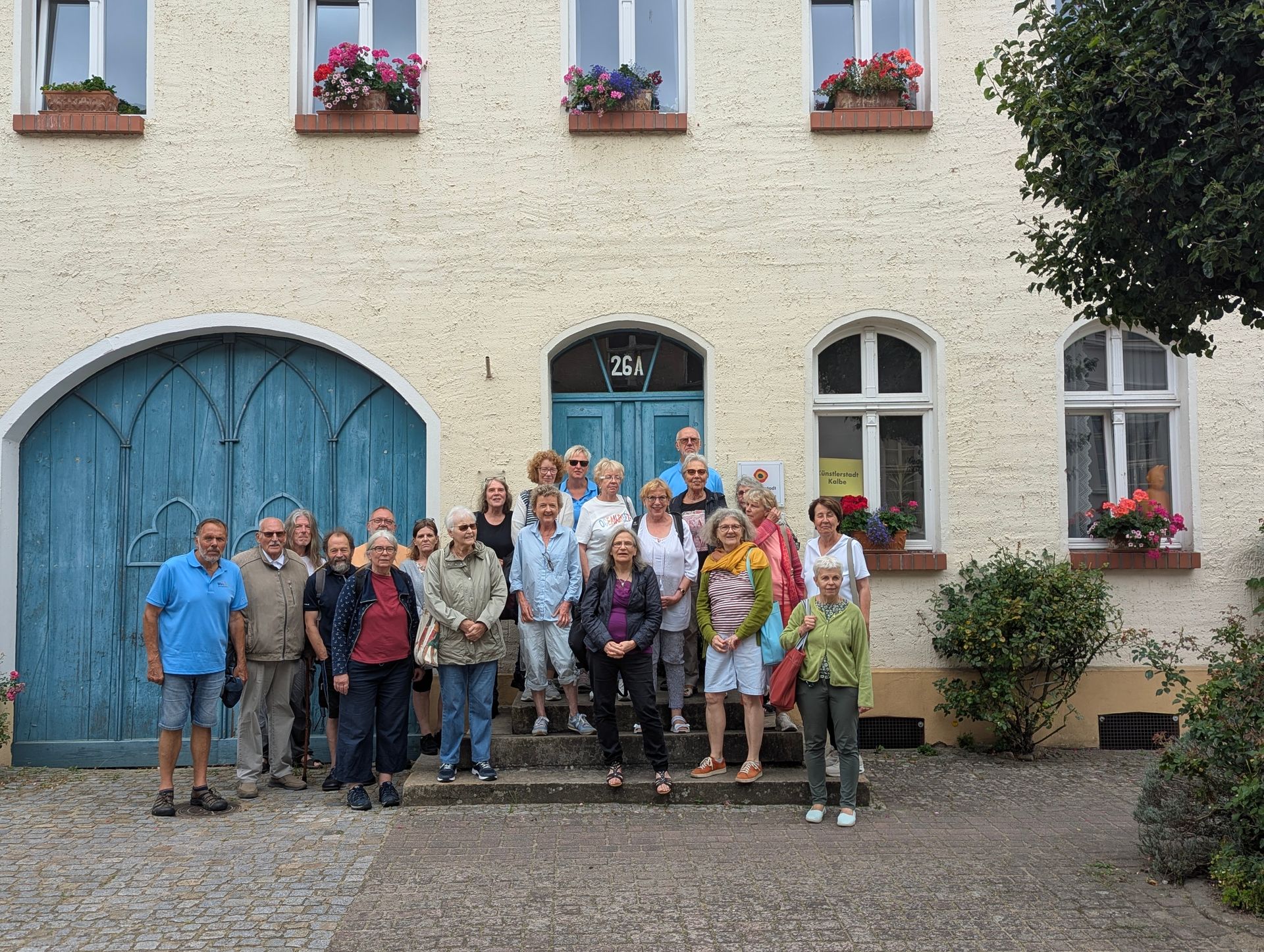Eine Gruppe von Menschen steht auf Stufen vor einem hellen Gebäude mit blauen Türen und Blumenkästen.
