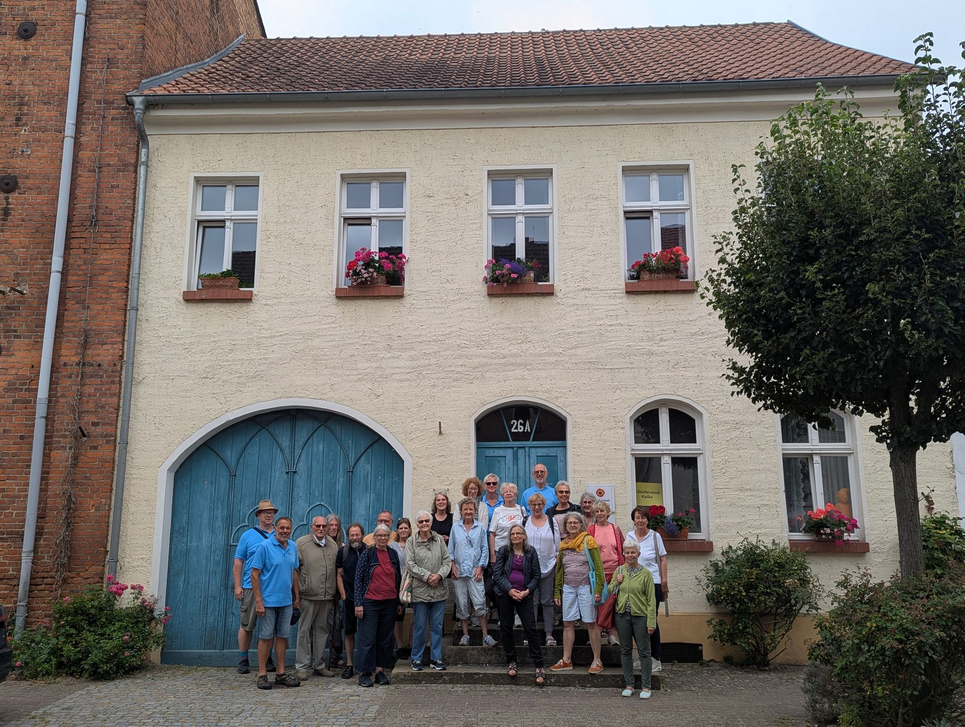 Eine Gruppe von Menschen steht vor einem hellgelben Gebäude mit blauer Tür und Fenstern mit Blumenkästen.