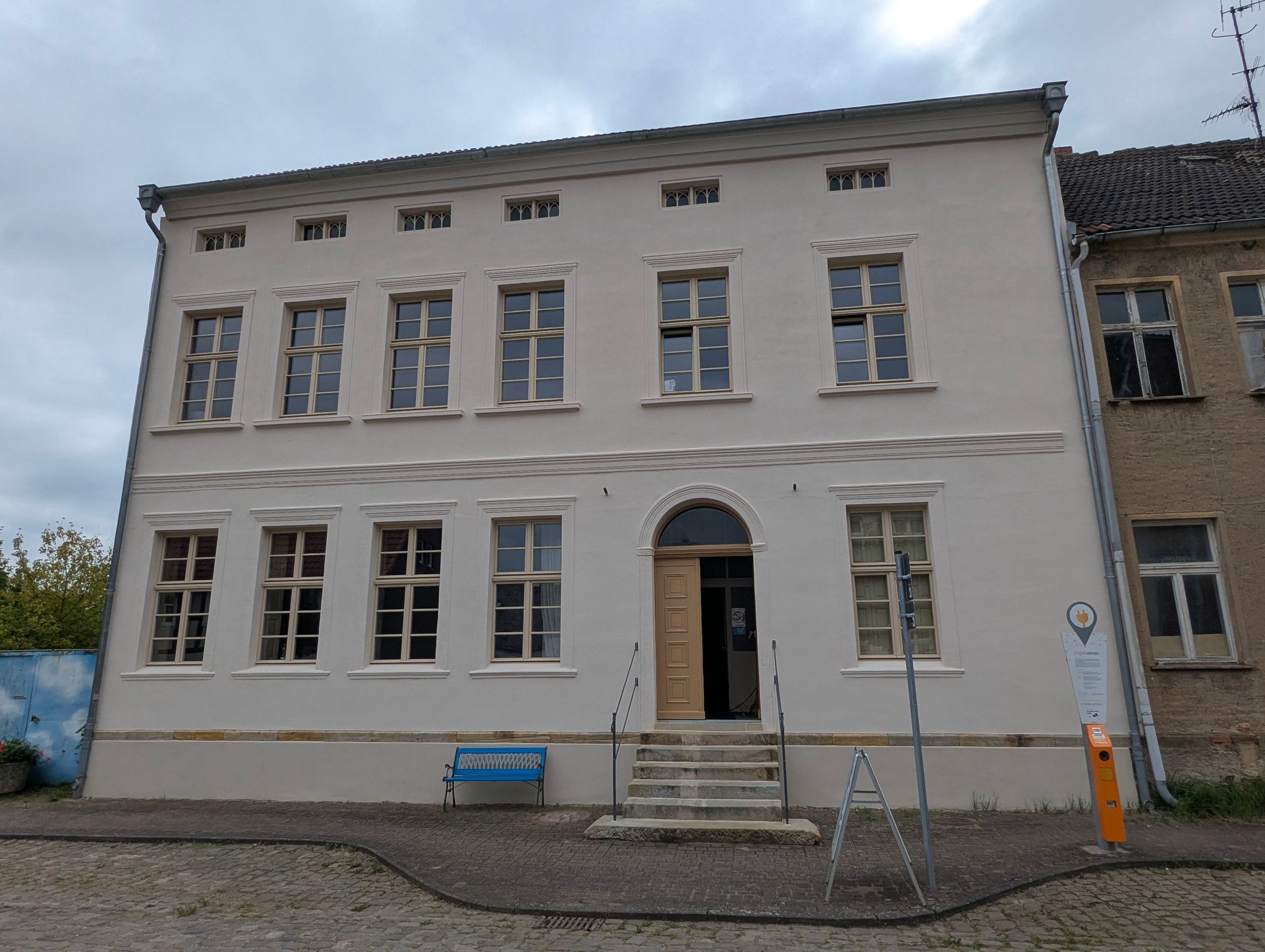 Zweigeschossiges, helles Gebäude mit vielen Fenstern. Offener, gewölbter Eingang mit Treppe. Bewölkter Himmel.