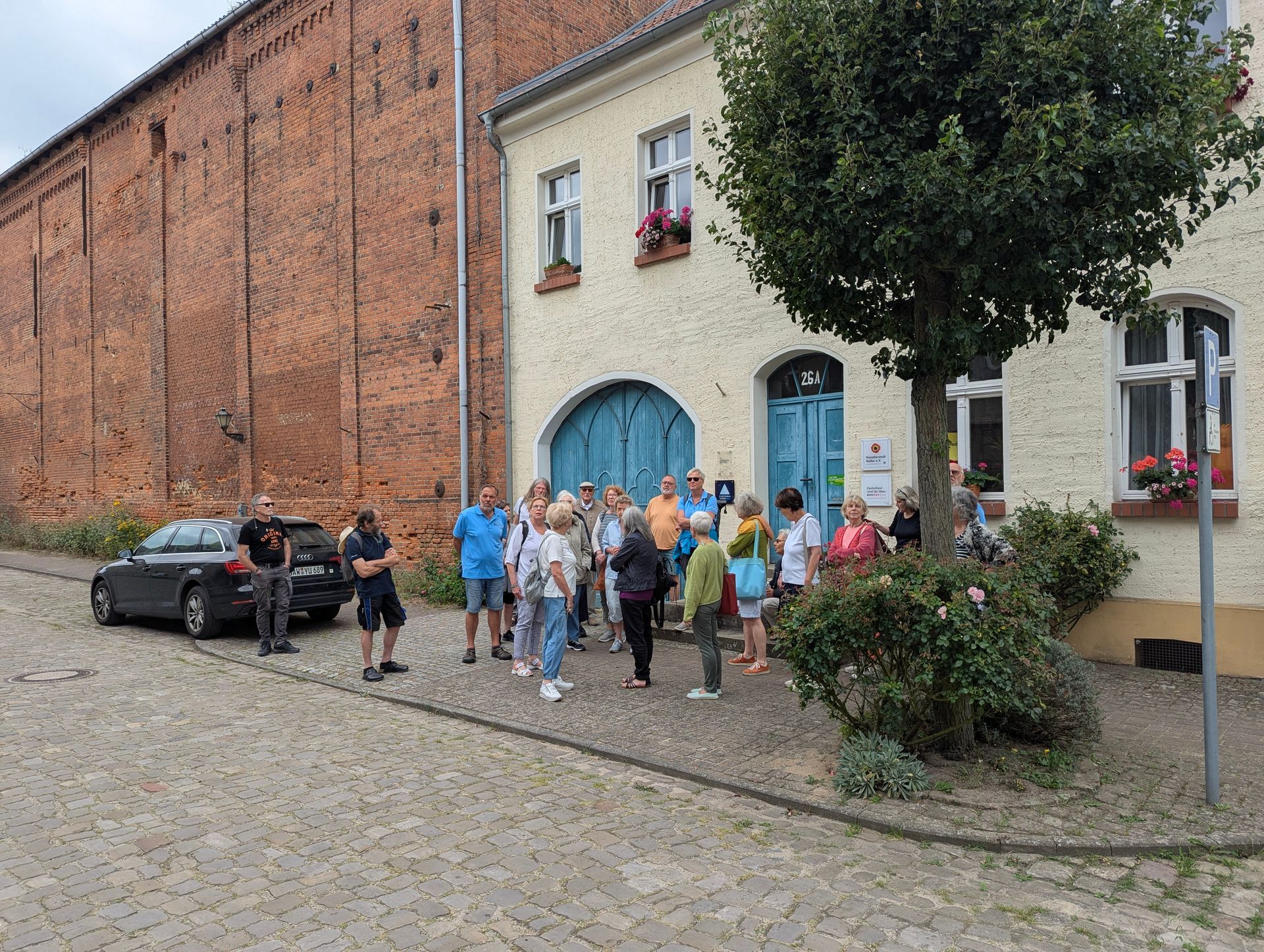 Eine Gruppe von Menschen vor einem Gebäude mit Backstein- und Stuckwänden, einige in der Nähe eines Baumes, andere bei einem Auto.