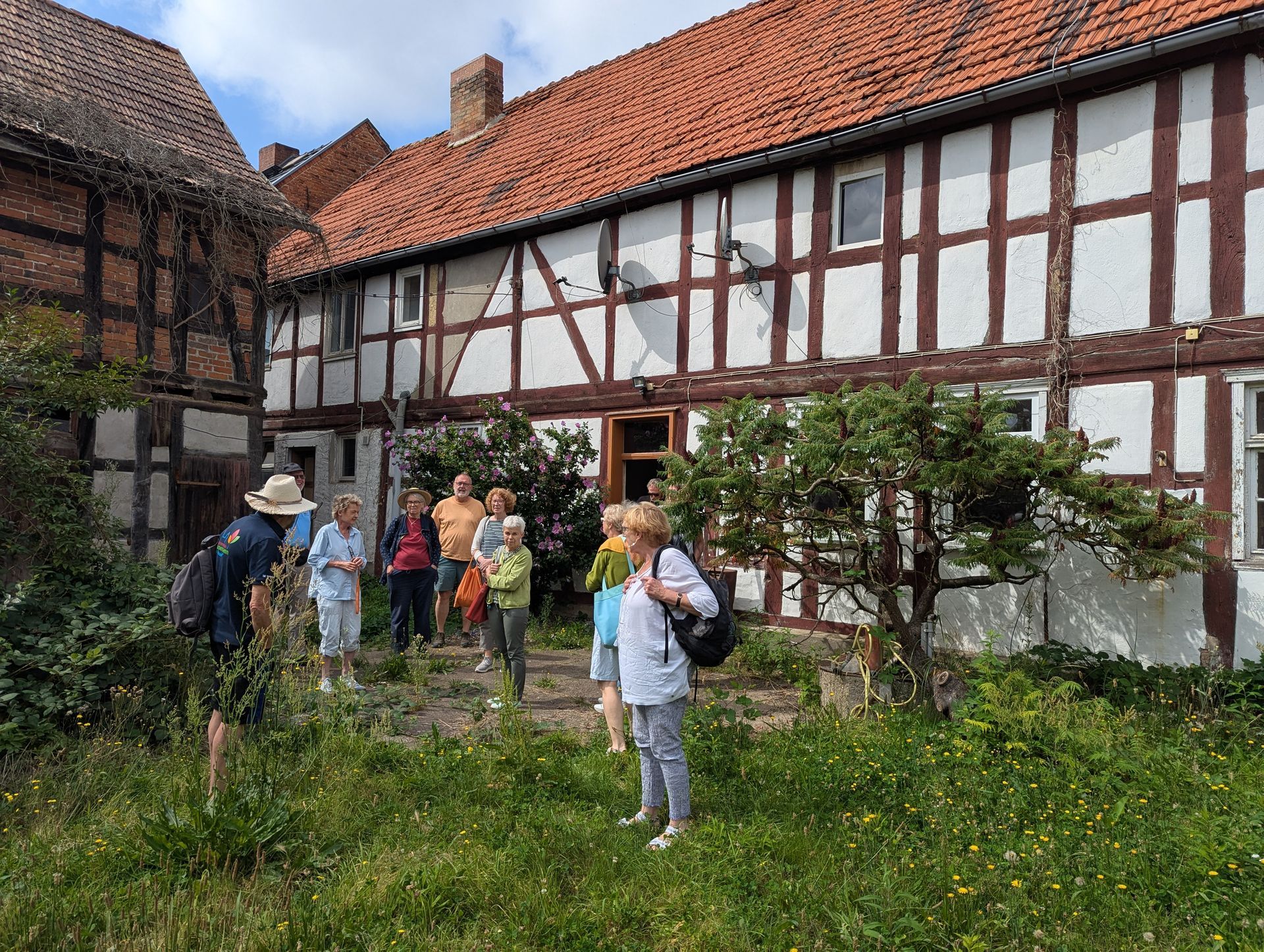 Eine Gruppe von Menschen steht vor einem Fachwerkhaus mit rotem Ziegeldach; im Vordergrund ist Grün zu sehen.