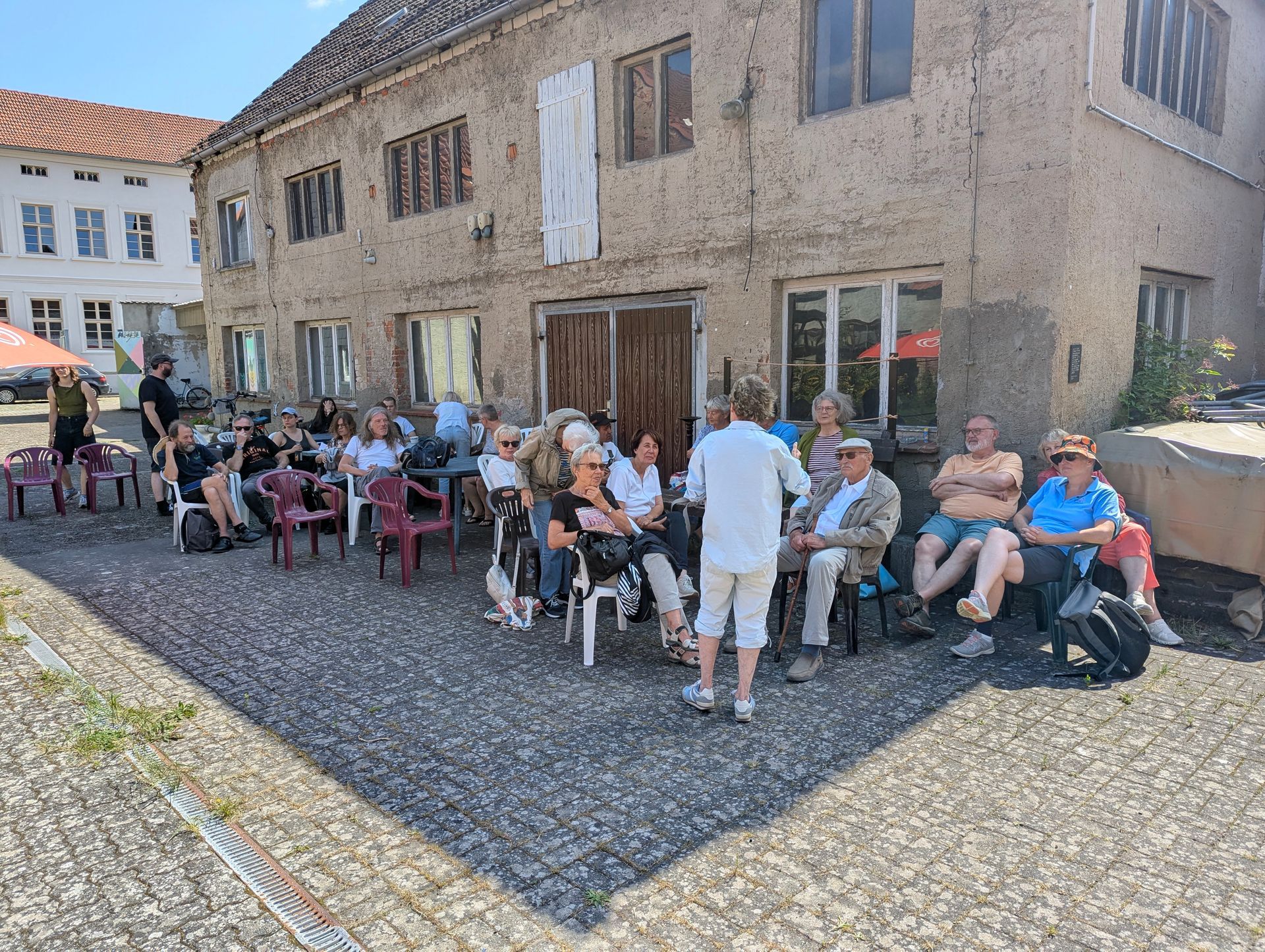 Eine Gruppe von Menschen sitzt im Freien und hört einem Mann vor einem verwitterten Gebäude zu.