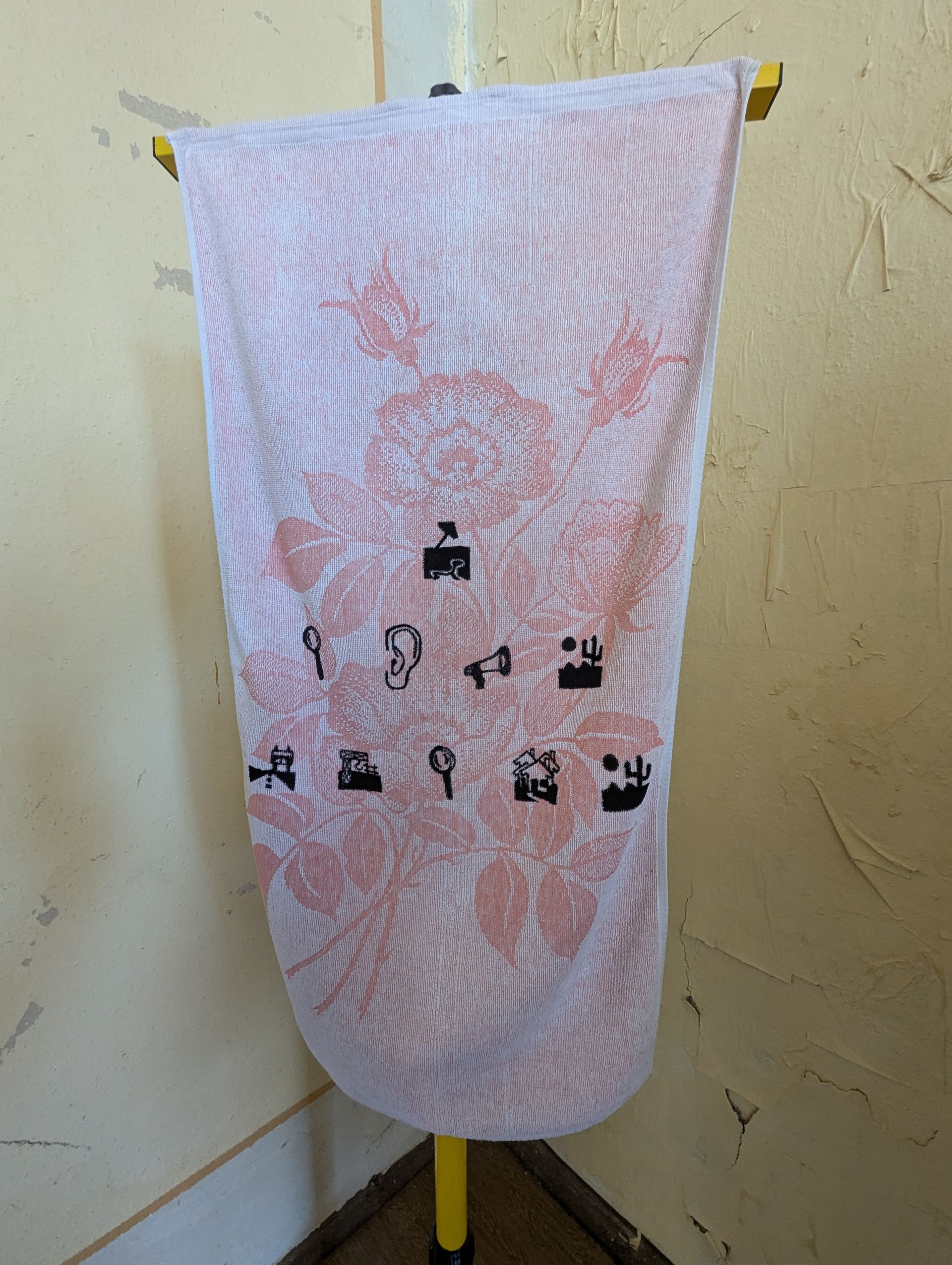 Banner mit rosa Muster und schwarzen Symbolen, das an einer gelben Stange vor einer beigen Wand hängt.