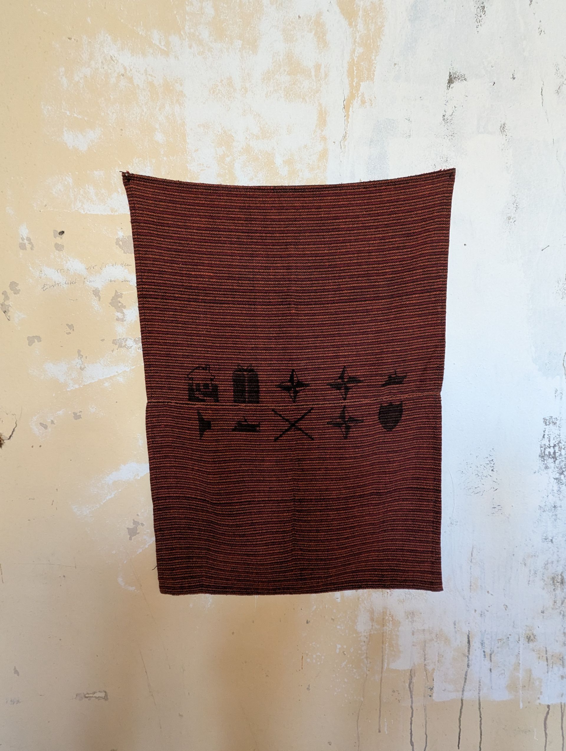 Gewebter brauner Stoff hing an einer strukturierten, hellen Wand; das Muster ähnelte einer Holzmaserung, am unteren Rand befand sich etwas Text.