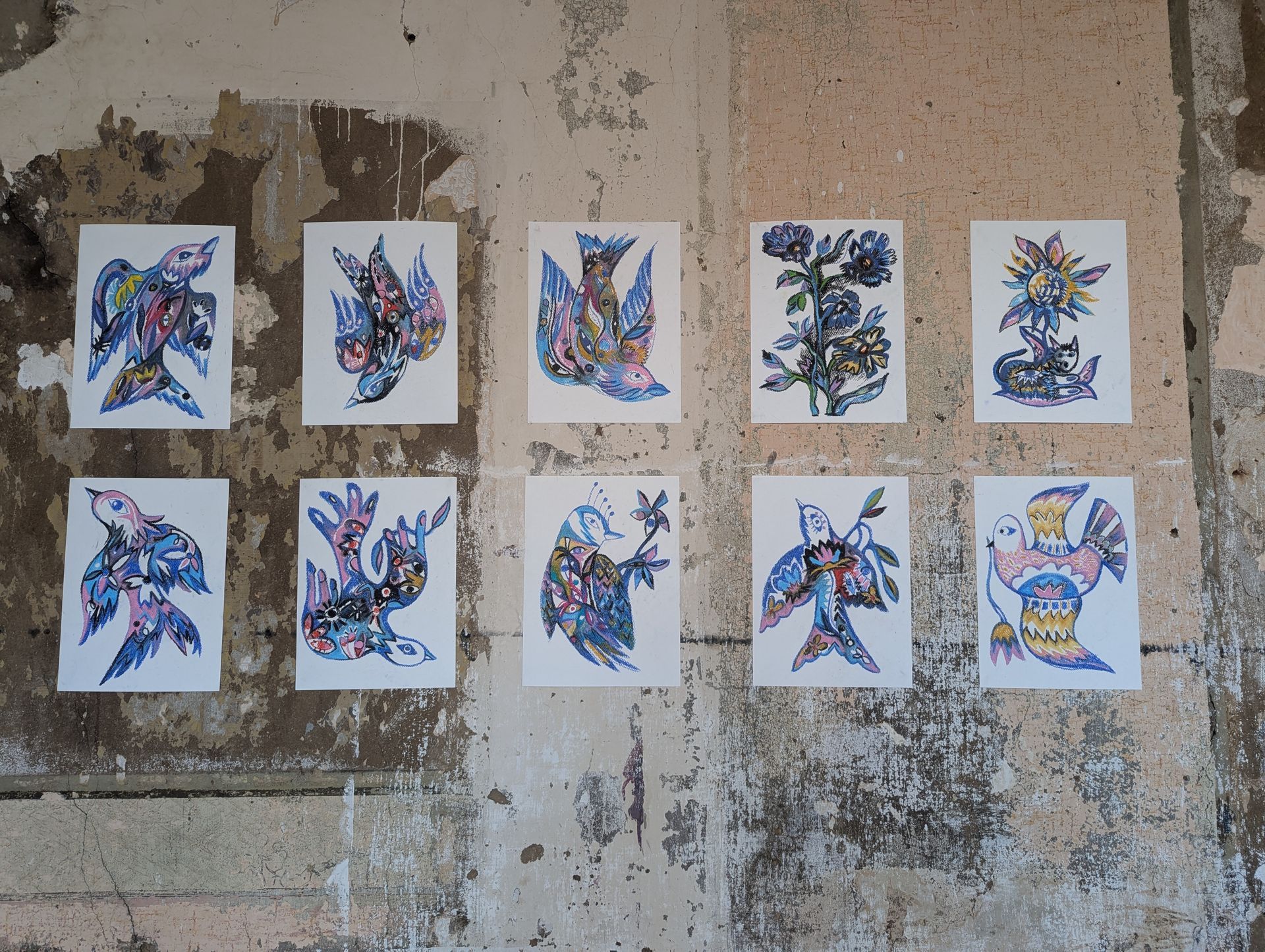Neun farbenfrohe, vogelähnliche Kunstwerke hängen an einer verwitterten, strukturierten Wand.
