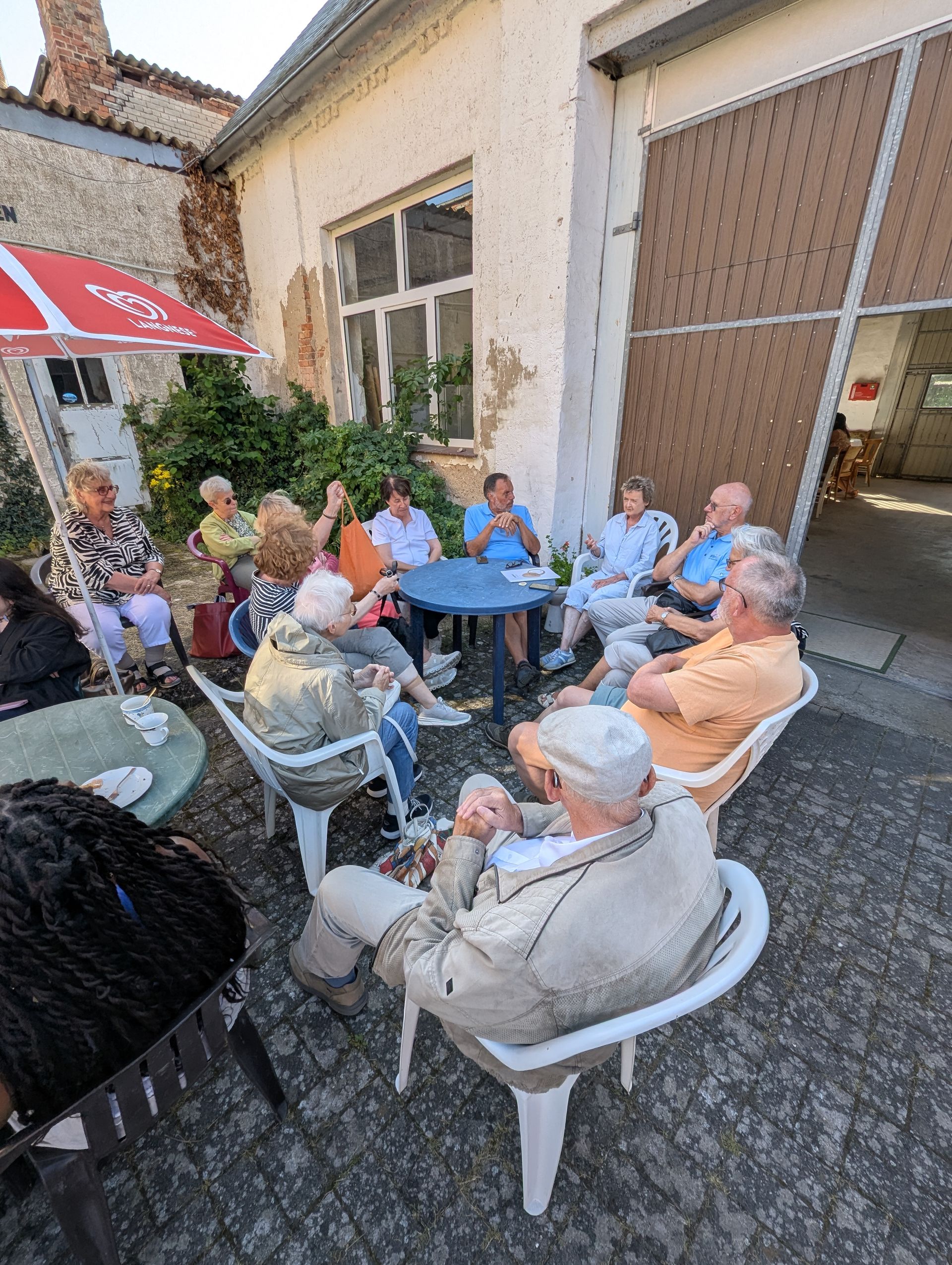 Eine Gruppe von Menschen sitzt im Freien um einen Tisch und unterhält sich. Ein roter Sonnenschirm spendet Schatten.