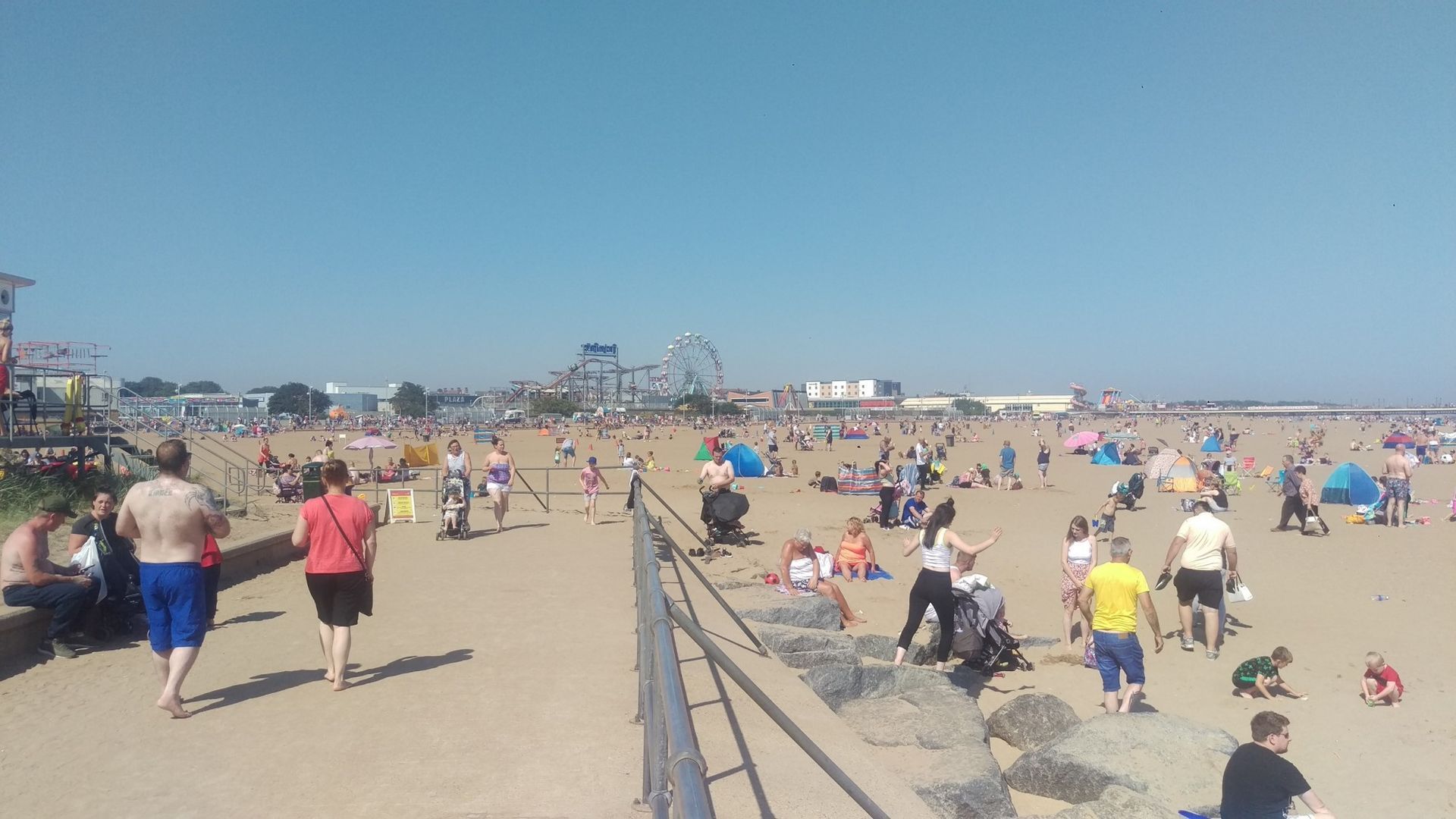 Strandszene mit vielen Menschen im Sand, Fahrgeschäfte eines Vergnügungsparks in der Ferne, sonniger Tag.