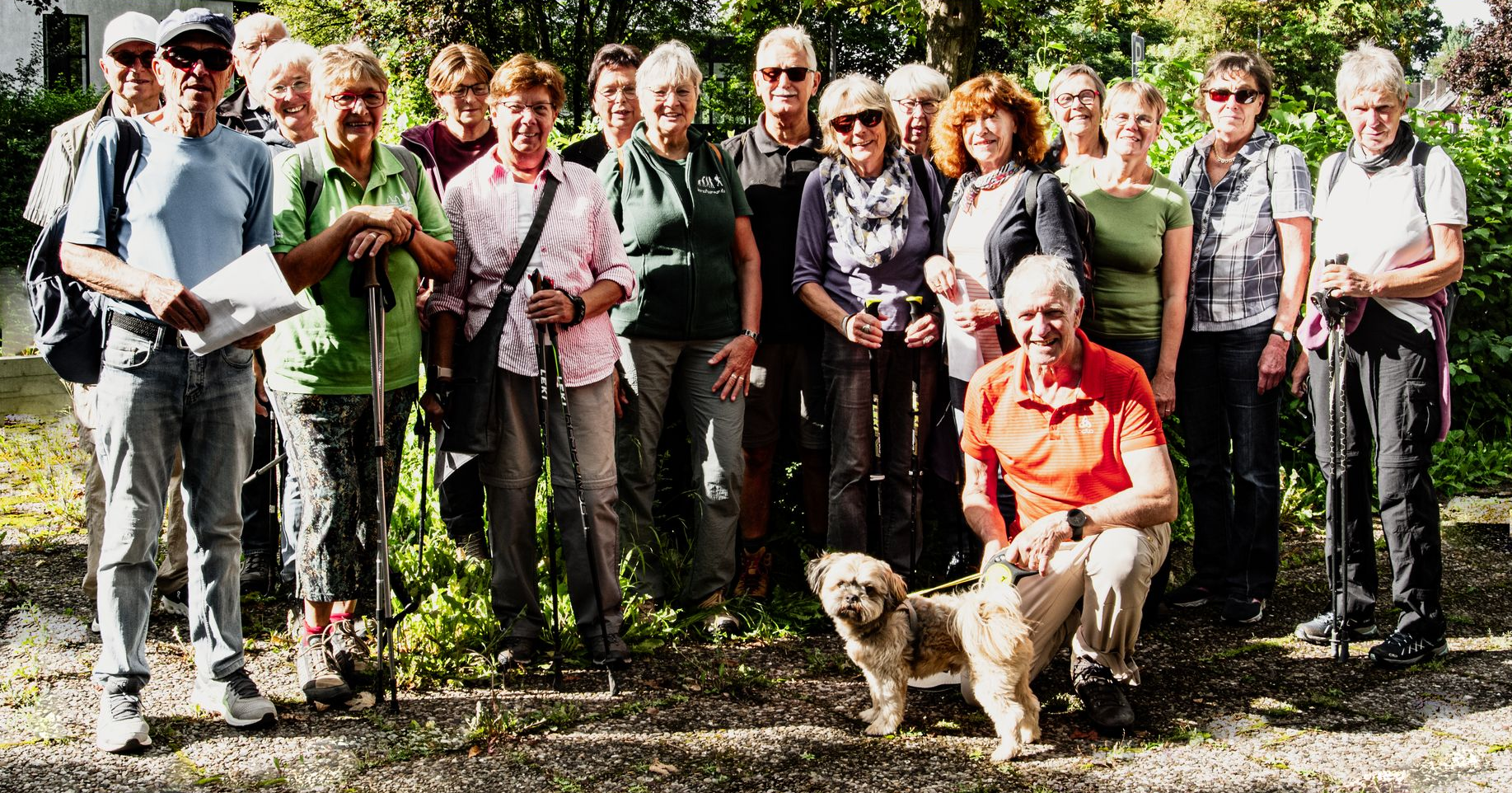 Eine Gruppe von Menschen im Freien, einige lächeln, mit einem kleinen Hund. Sonniger Tag, im Hintergrund Grün.