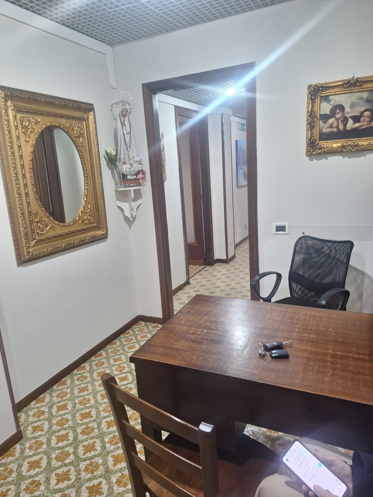 Piccolo ufficio con scrivania e sedia in legno, specchio a parete, quadri incorniciati e porta aperta in una stanza piastrellata.