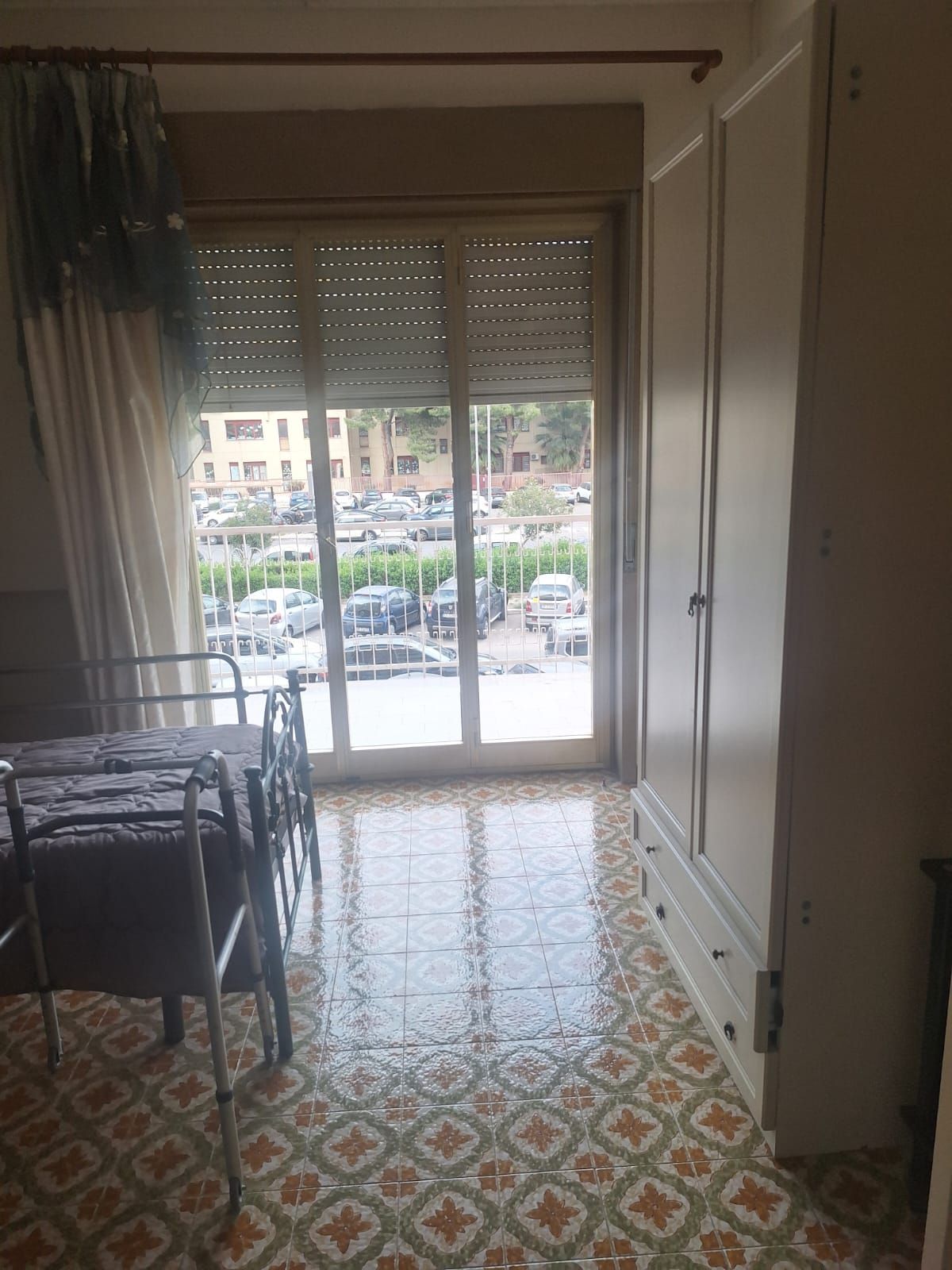 Piccola stanza con pavimento piastrellato a motivi geometrici, letto e porte finestre che si aprono su una vista sulla strada.