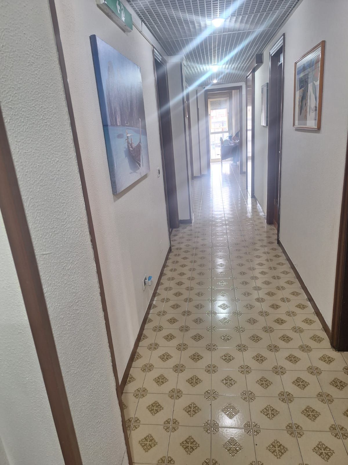 Stretto corridoio con pavimento piastrellato decorato, quadri incorniciati alle pareti e luce intensa in fondo.