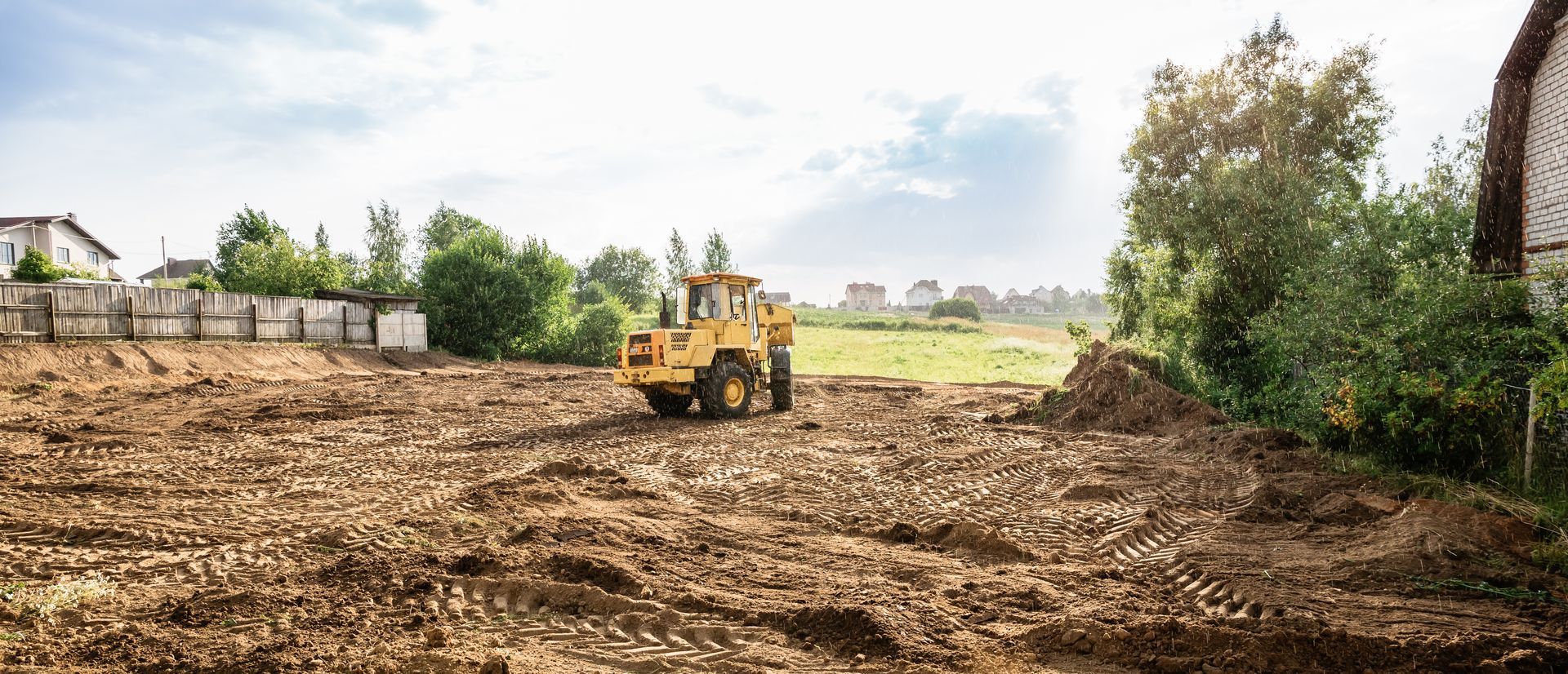 Bulldozer leveling dirt on a cleared construction site with trees and houses nearby.