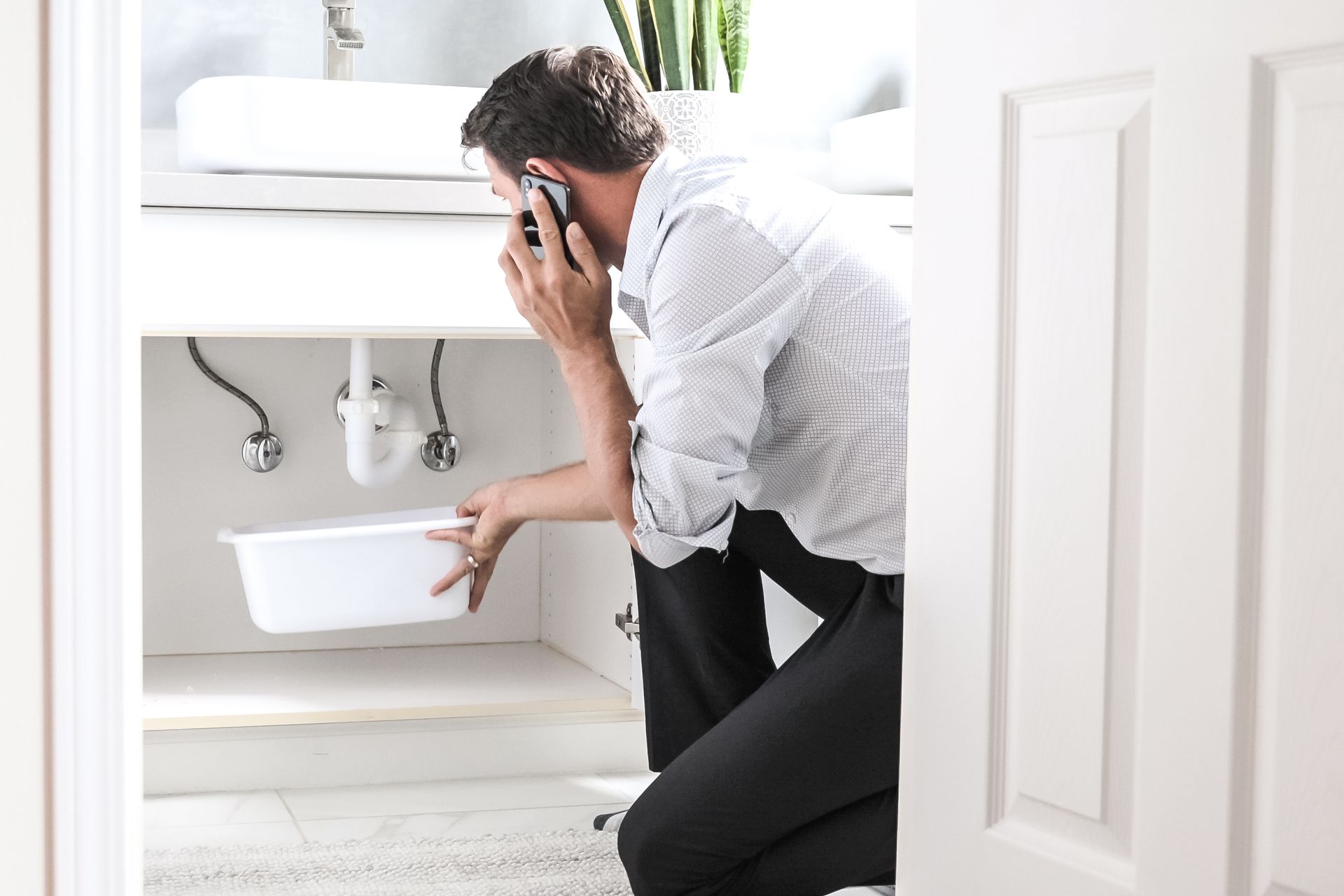Man crouching under bathroom sink, holding phone and inspecting a white basin.