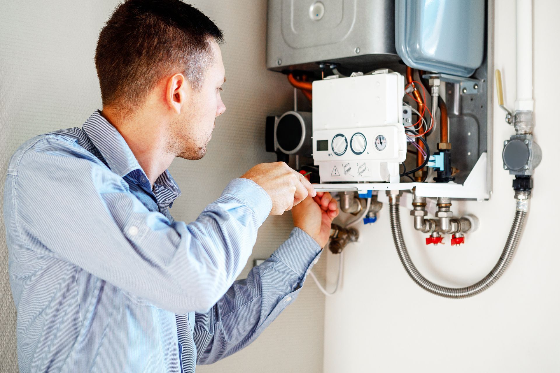 Technician adjusting controls on a wall-mounted boiler with exposed pipes and hoses