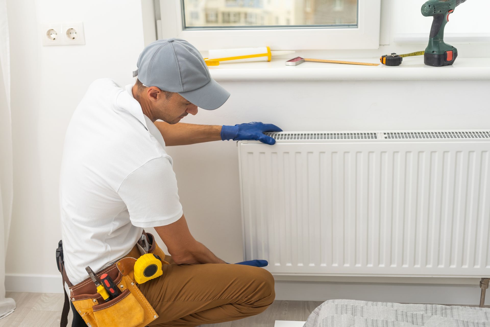 Technician inspecting a white radiator in a bright room, wearing a cap, gloves, and tool belt.