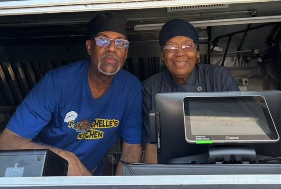 Two smiling vendors behind a food truck counter with a cash register in front of them