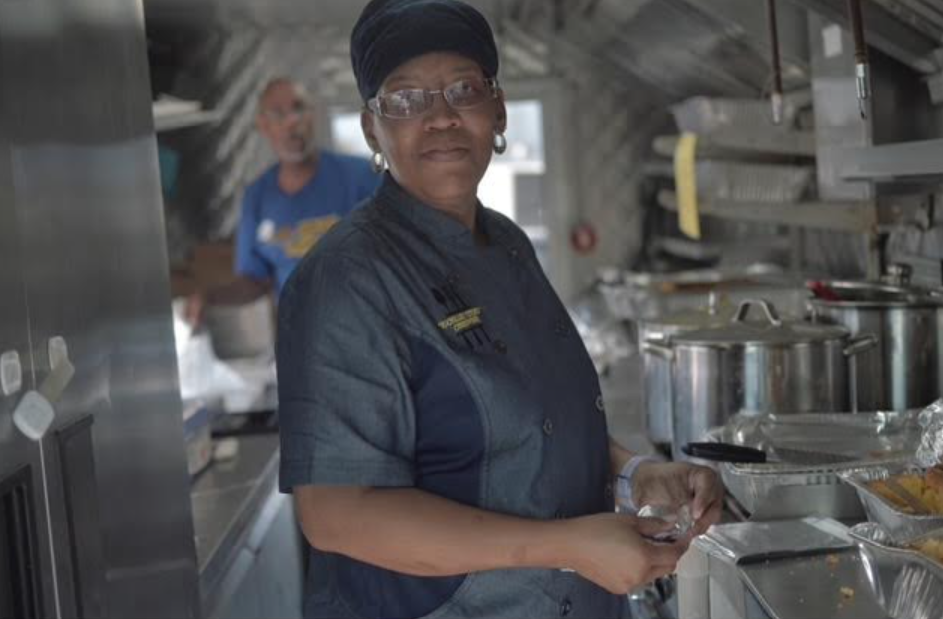 Chef smiling in a stainless-steel kitchen, holding a utensil beside trays of prepared food.