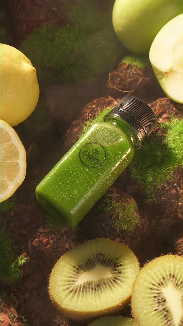 Green juice bottle surrounded by kiwi, lemon, and green apples on a mossy surface.