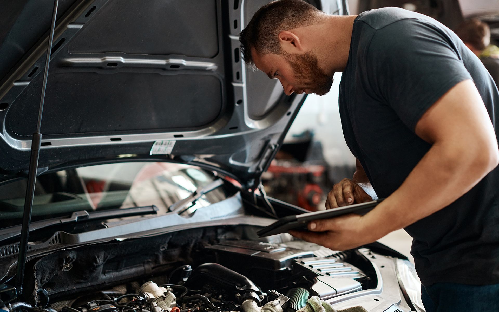 Mechanic inspecting car engine, using tablet in auto repair shop.