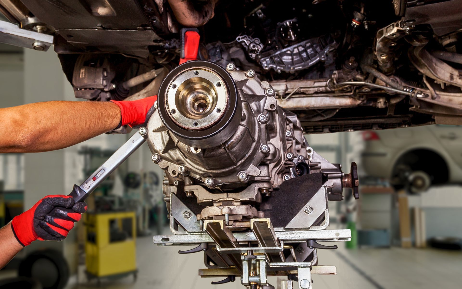 Mechanic removing a car's differential with a wrench, suspended on a lift. Red gloves and workshop setting.