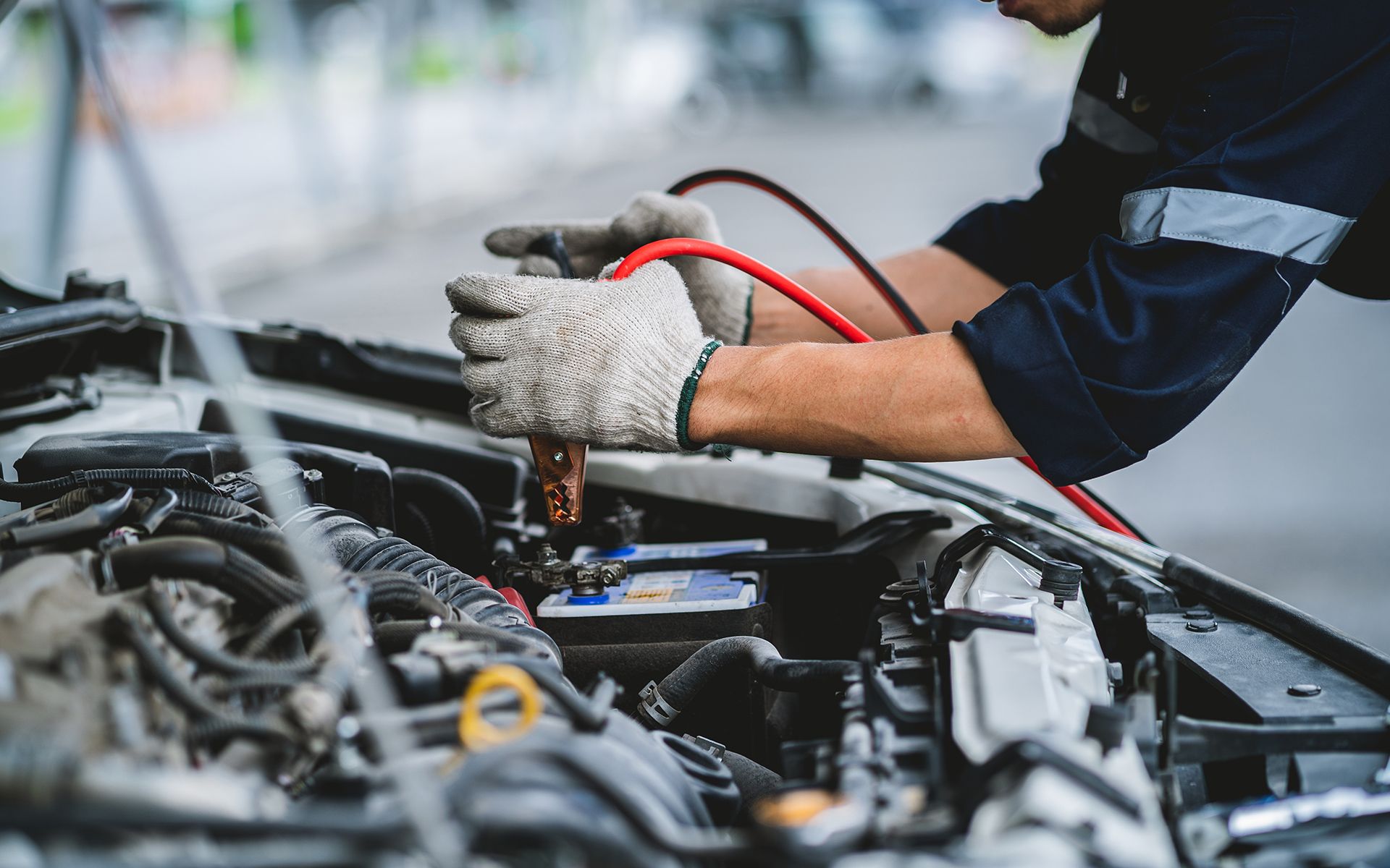 Mechanic in gloves connecting jumper cables to a car battery in an engine bay.
