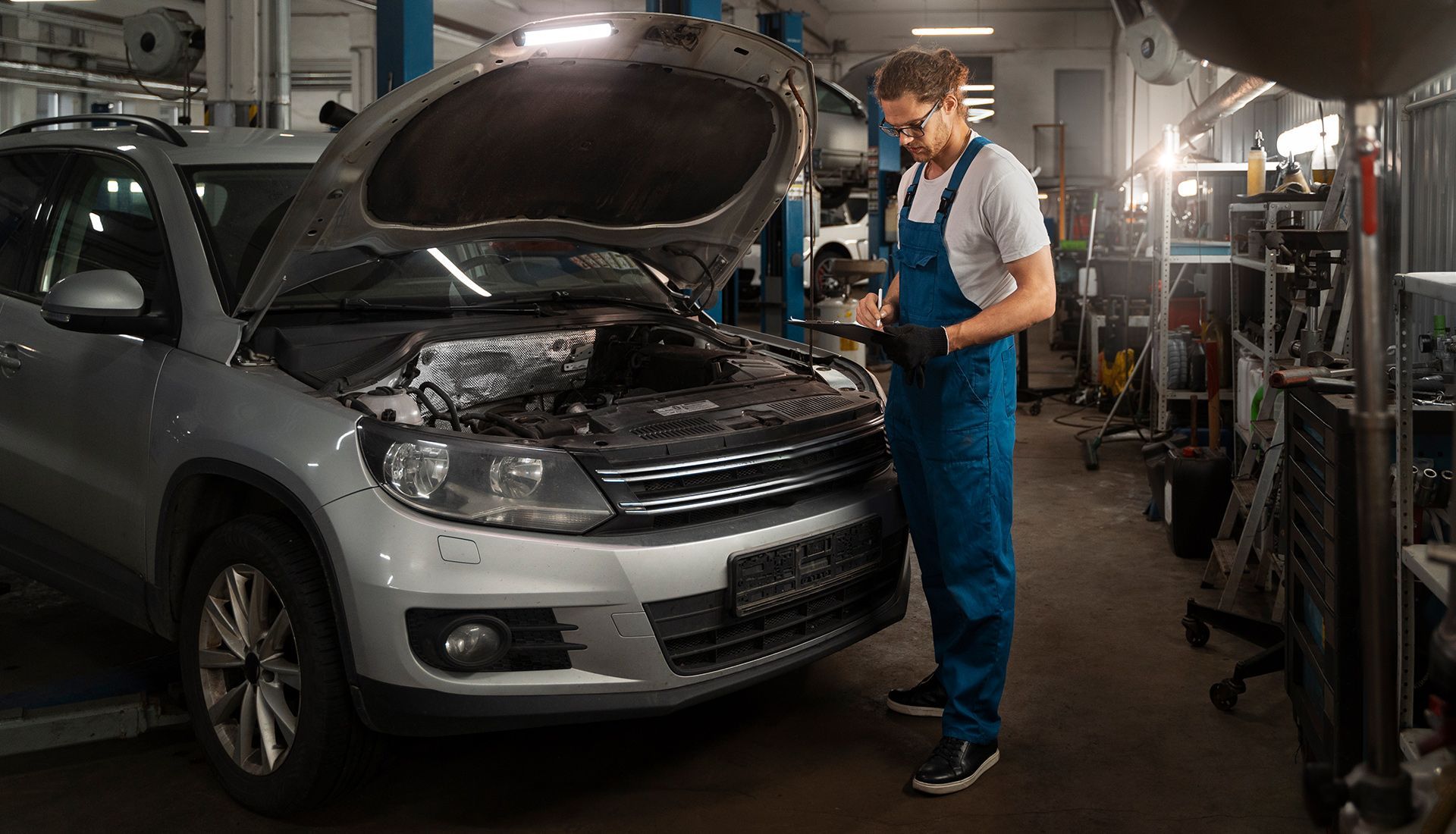 Mechanic in blue overalls inspecting a silver car with the hood open in a garage.